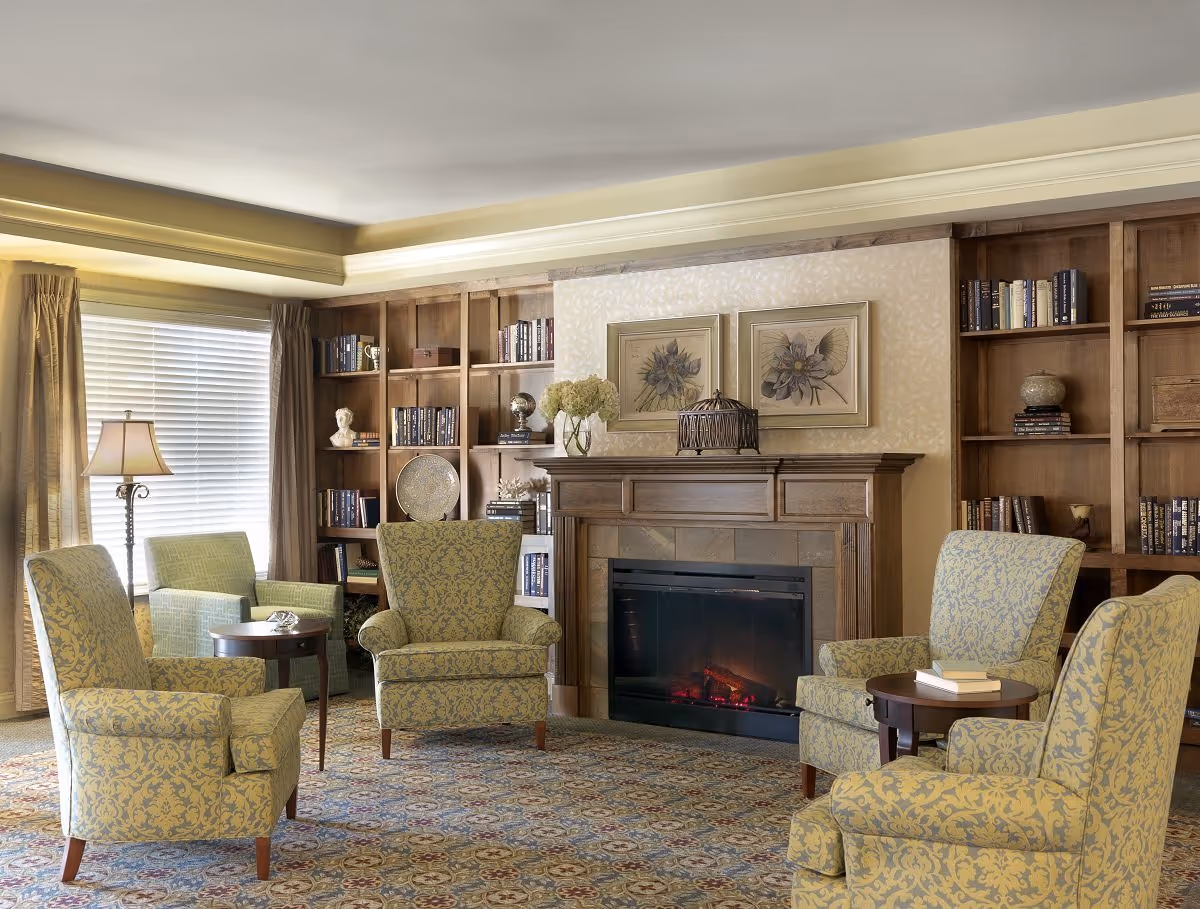 A cozy living room area with four patterned armchairs arranged around two small round wooden tables. There is a fireplace with a wooden mantel, decorated with framed floral artwork and a vase of flowers. Built-in wooden bookshelves filled with books and decorative items flank the fireplace. A window with closed blinds and beige curtains allows natural light into the room.