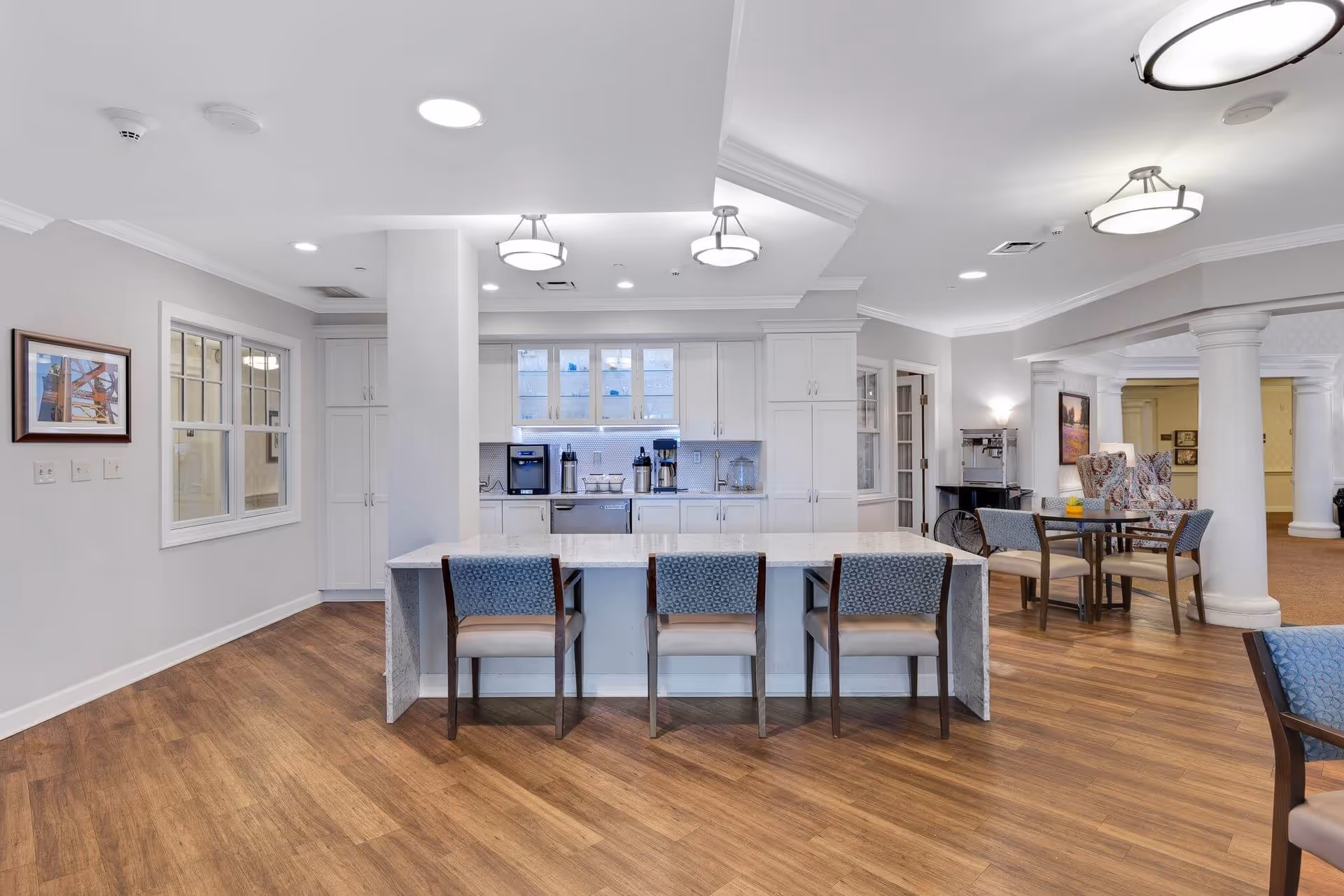 Open communal kitchen and seating area with a marble island, stools, cabinets, and adjacent dining tables.