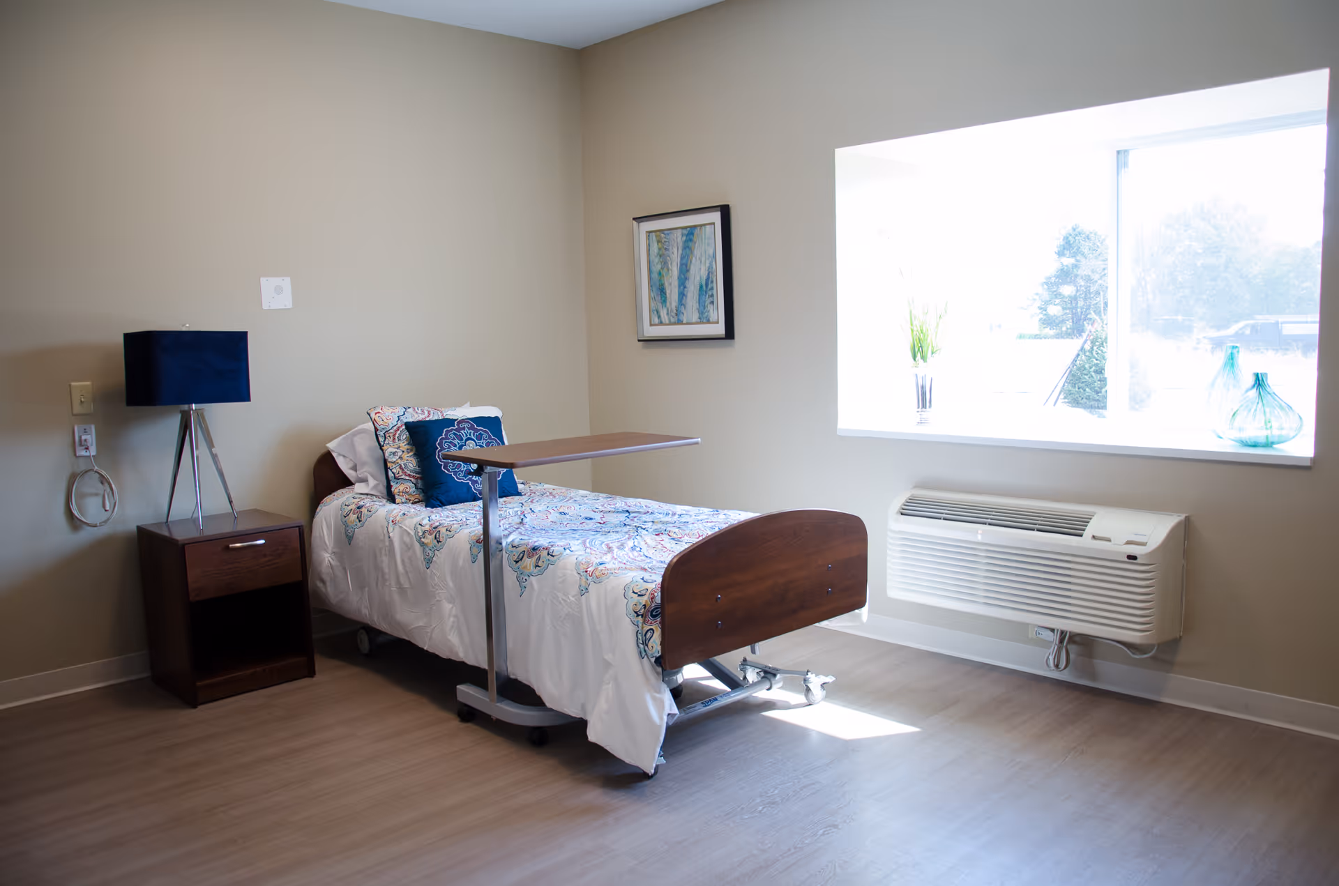 A simple, clean room with a single hospital-style bed covered in a patterned bedspread and pillows. Next to the bed is a wooden nightstand with a blue lamp. A large window lets in natural light, with decorative vases on the windowsill. There is a wall-mounted air conditioning unit below the window and a framed artwork on the wall.