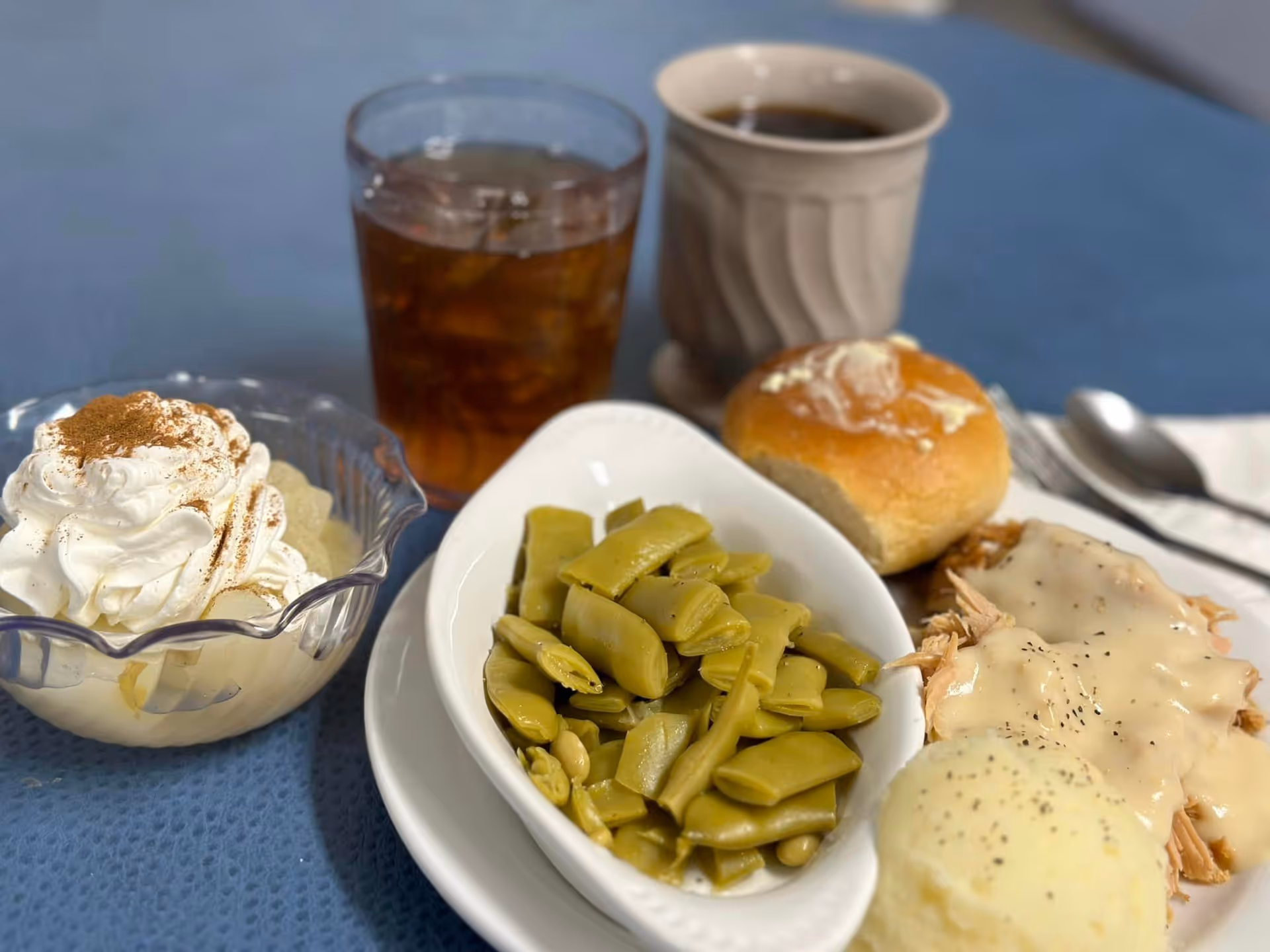 A meal served on a white plate featuring cooked green beans, mashed potatoes with gravy, and shredded turkey with gravy. There is a buttered dinner roll next to the plate. A glass of iced tea and a cup of coffee are placed behind the plate. A dessert bowl with whipped cream and a sprinkle of cinnamon is also visible on the side, all set on a blue tablecloth.