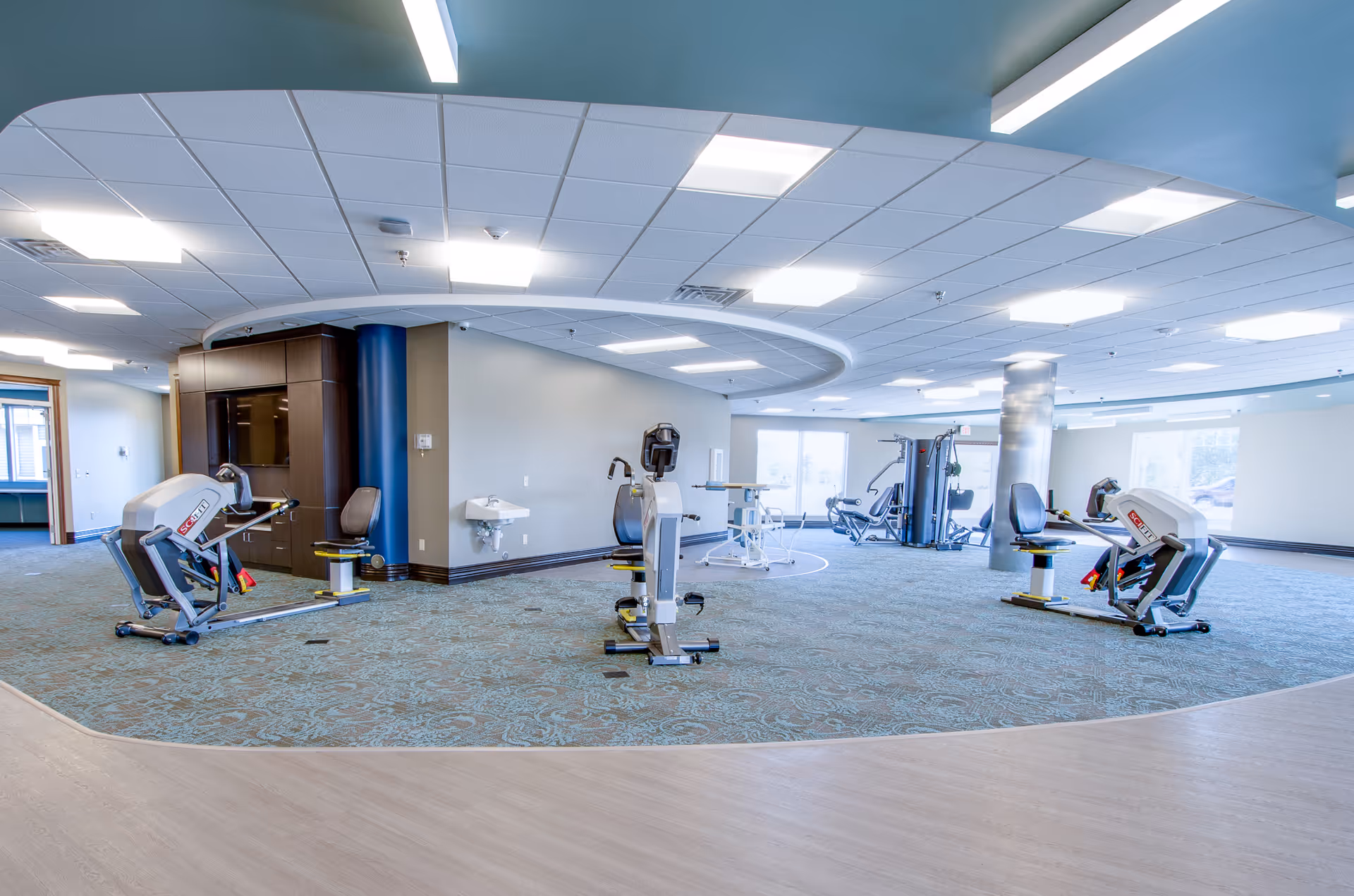 A spacious rehabilitation exercise room with various exercise machines including seated leg press and multi-functional gym equipment. The room has a patterned carpeted floor, large windows allowing natural light, and a ceiling with recessed lighting. There is a small sink mounted on the wall and a wooden cabinet with a TV screen.
