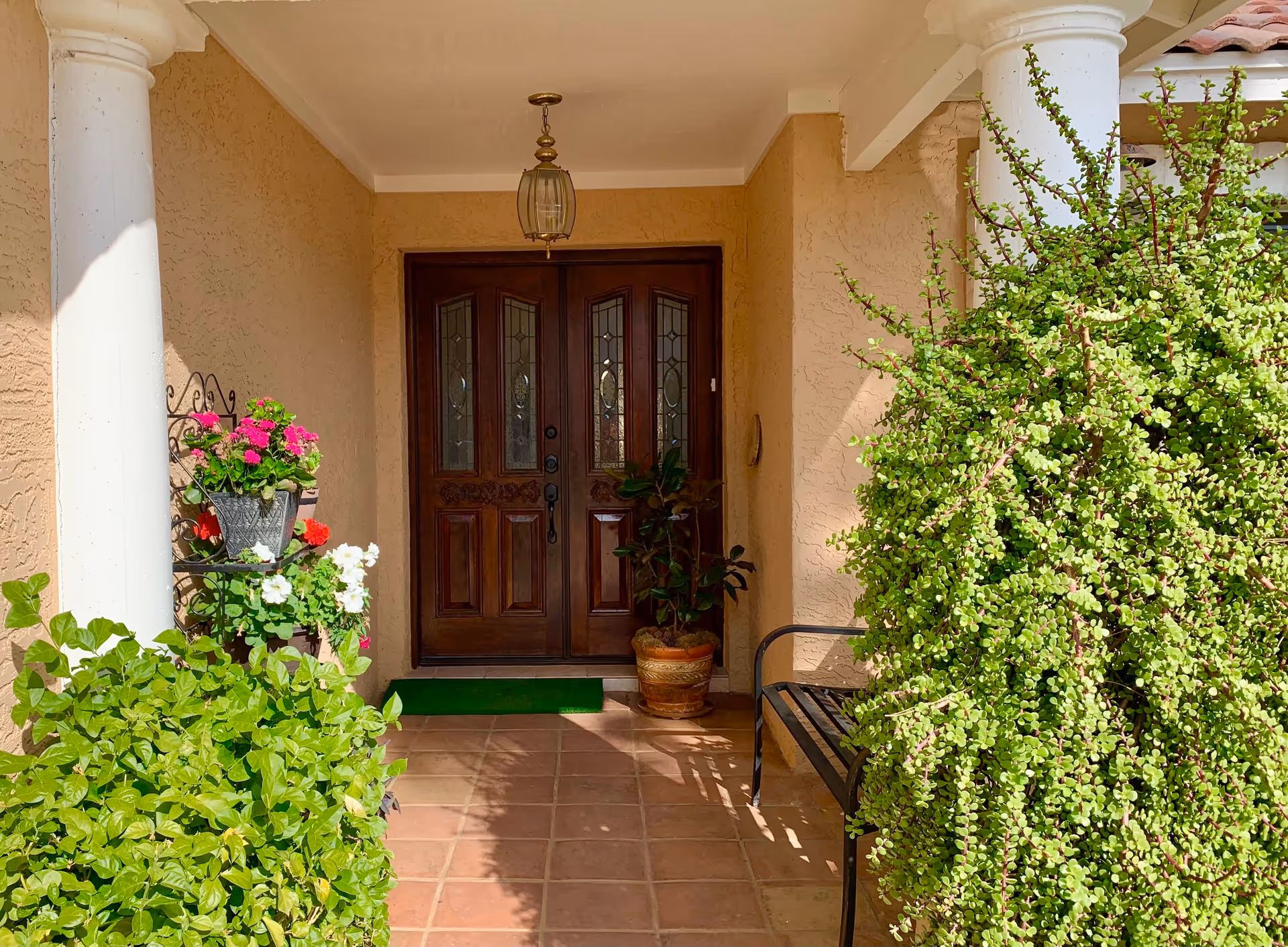 Front entrance of a building with double wooden doors featuring decorative glass panels, surrounded by beige stucco walls. There are green plants and flowers on either side of the entrance, a black metal bench on the right, and a hanging lantern light above the doorway.