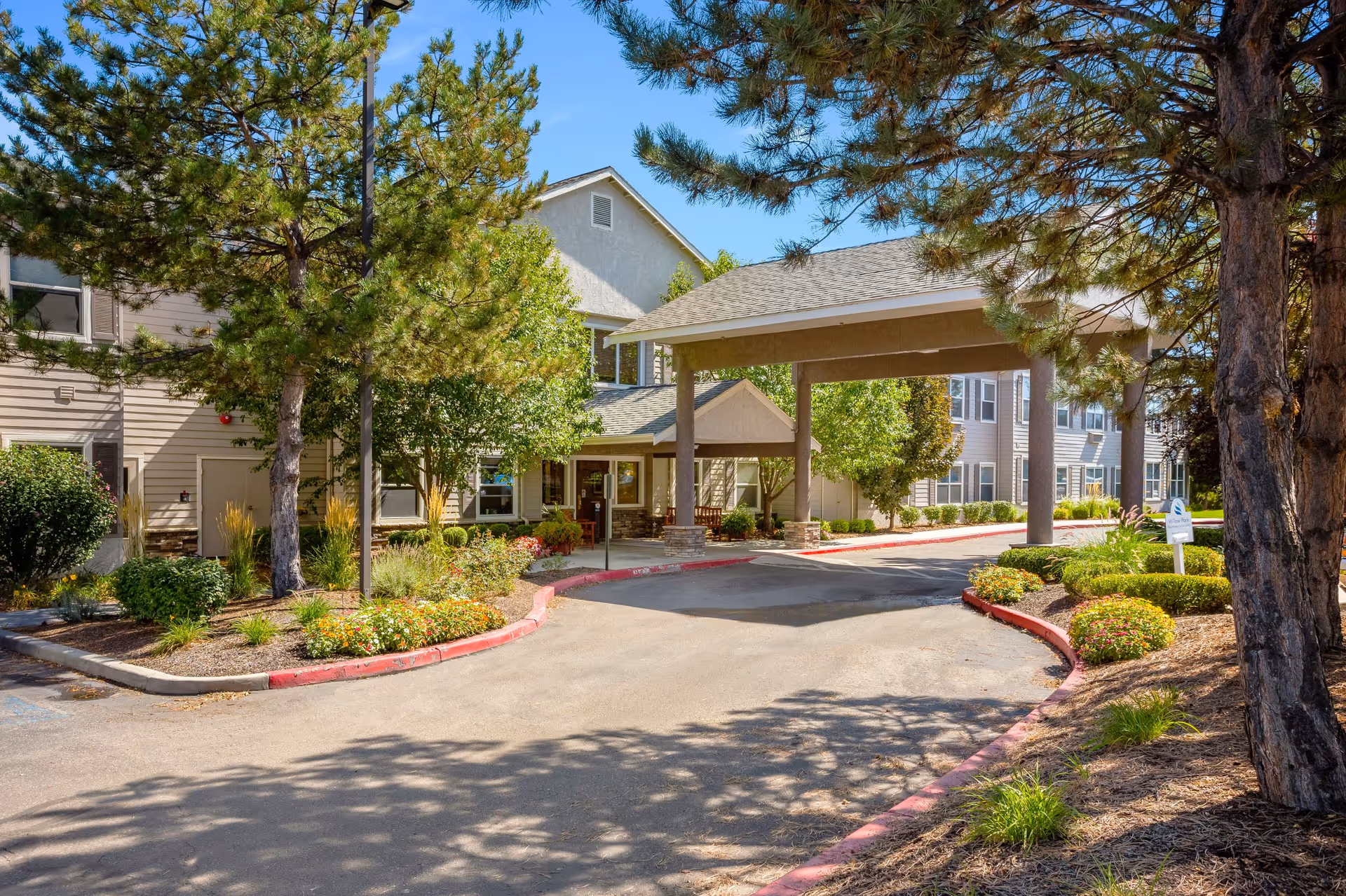 Entrance driveway to a senior living facility with a covered drop-off area, surrounded by trees, shrubs, and landscaped flower beds under a clear blue sky.