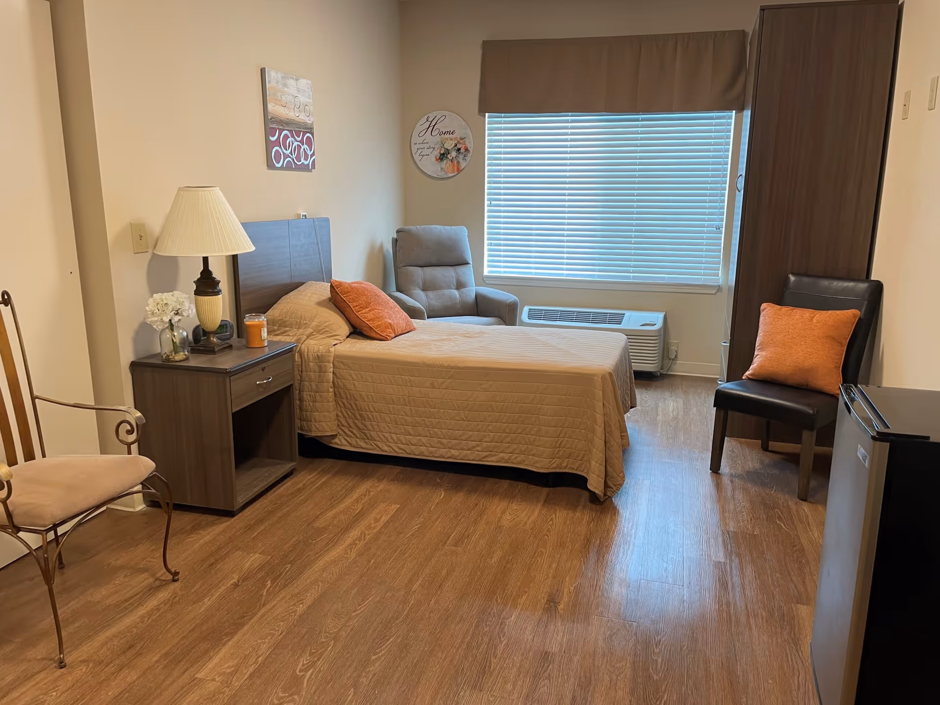 A cozy bedroom with a single bed covered in a beige quilt and an orange pillow. Next to the bed is a wooden nightstand with a lamp, a small vase with white flowers, and a candle. There is a cushioned armchair near a window with closed blinds, and a wooden wardrobe stands against the wall. A black chair with an orange pillow is placed near a small refrigerator. The room has wooden flooring and light-colored walls with two decorative wall hangings.