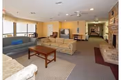 Bright assisted-living common room with sofas, a coffee table, fireplace, TV and a long corridor.