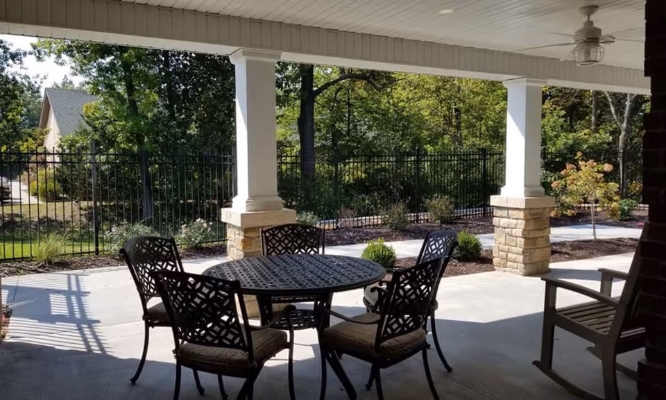 Covered outdoor patio area with a round metal table and four matching chairs with cushions. The patio has white columns with stone bases and overlooks a fenced garden area with trees and shrubs.