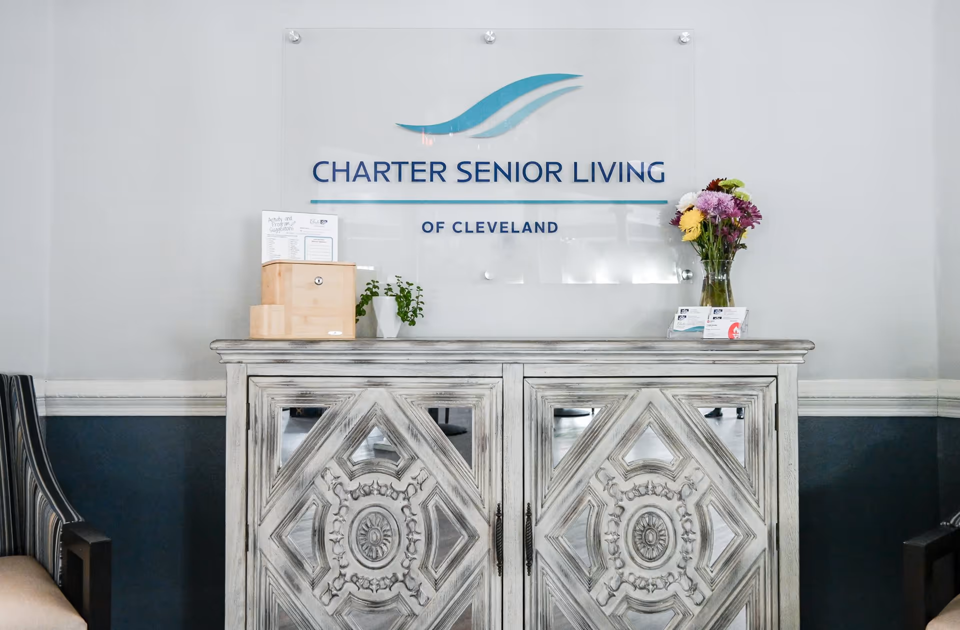 Decorative wooden cabinet in a lobby beneath a wall sign reading 'Charter Senior Living of Cleveland' with a vase of flowers on top.