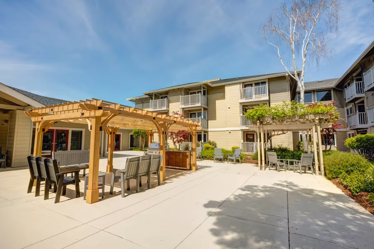 Outdoor courtyard area of a senior living facility with wooden pergolas, tables, and chairs arranged for seating and socializing. The building surrounding the courtyard is three stories high with balconies and windows. The sky is clear and blue.