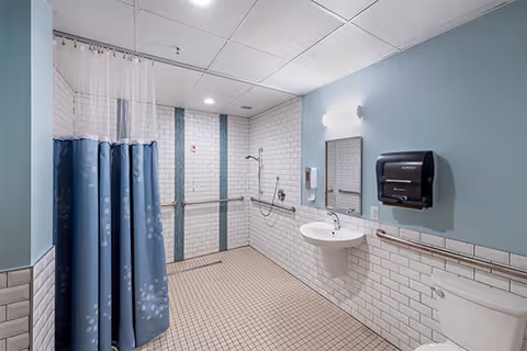 A clean, accessible bathroom with light blue walls and white subway tiles. The room features a walk-in shower area with a blue and white patterned shower curtain, a wall-mounted sink with a mirror above it, a paper towel dispenser, and a toilet. The floor is covered with small square beige tiles, and there are grab bars installed along the walls for support.