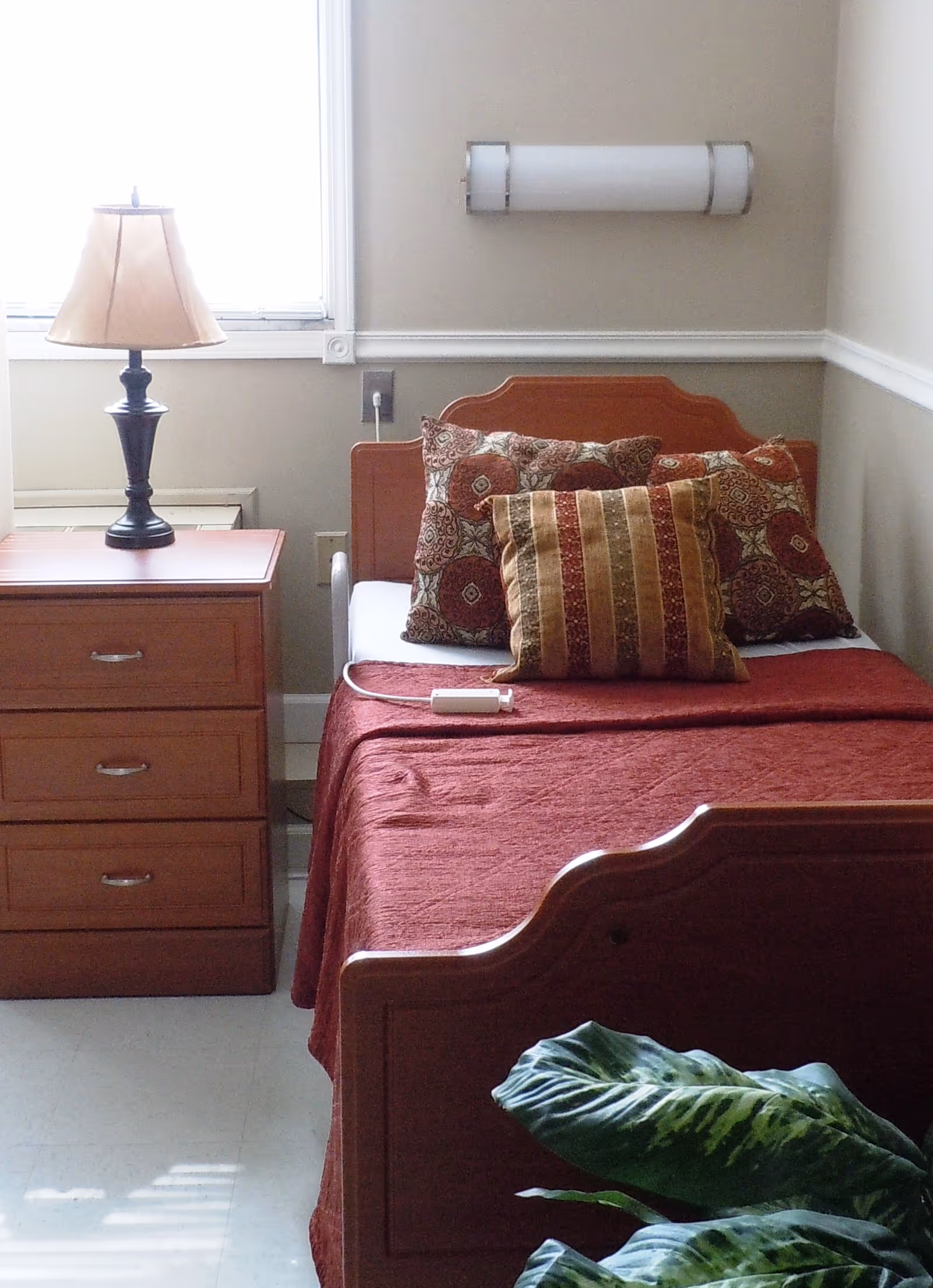 A small bedroom with a single wooden bed covered with a red bedspread and multiple decorative pillows. Next to the bed is a wooden nightstand with three drawers and a lamp on top. A window is visible behind the nightstand, and a green leafy plant is partially visible in the foreground.