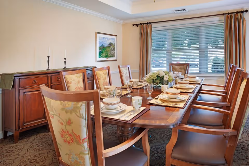 A formal dining room with a long wooden table set for eight people. The table is arranged with plates, bowls, cups, glasses, and silverware on placemats. There is a floral centerpiece in the middle of the table. The room has a large window with striped curtains, a wooden sideboard with two candlesticks, and a framed landscape painting on the wall.