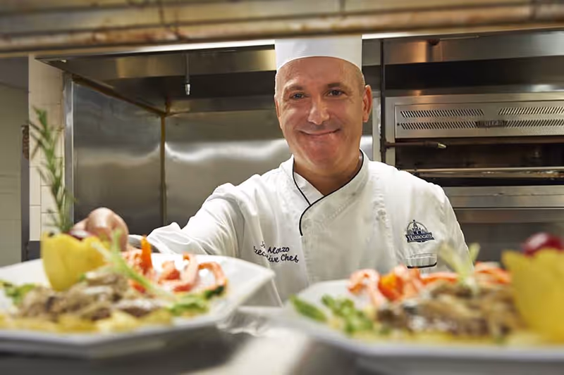 A smiling chef in a white uniform and hat stands in a commercial kitchen, presenting two plates of gourmet food with garnishes.