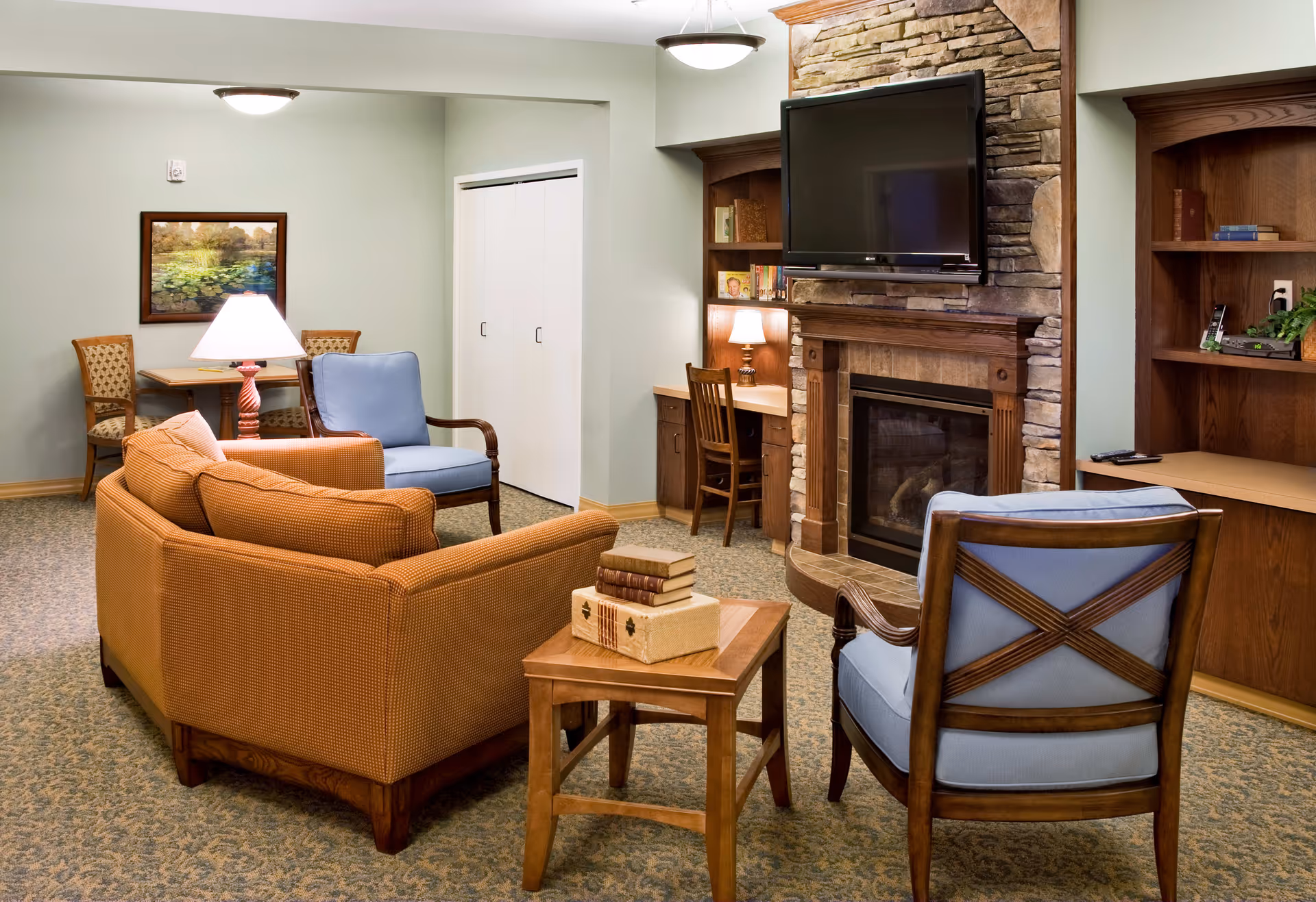 A cozy living room area in a senior living facility featuring a stone fireplace with a mounted flat-screen TV above it. The room has a brown sofa, two blue cushioned wooden armchairs, a small wooden side table with stacked books, and a wooden desk with a chair and a lamp. There is also a small dining table with two chairs and a framed painting on the wall. The carpet is patterned and the walls are painted light green.