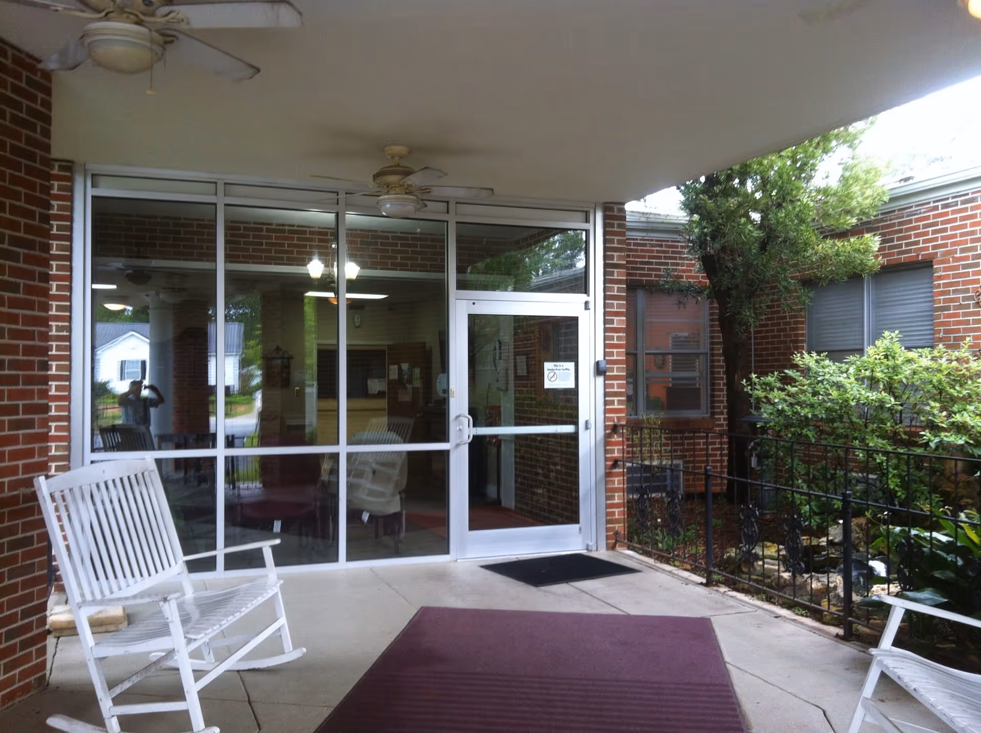 Covered outdoor entrance area of a brick building with glass doors and windows. There are two white rocking chairs on the concrete floor, a purple mat, ceiling fans, and some greenery on the right side near a black metal railing.