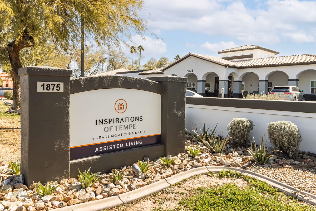 Exterior view of Inspirations Of Tempe assisted living facility showing the main entrance building with arches and a sign in front displaying the facility name and address number 1875, surrounded by desert landscaping with rocks and plants.