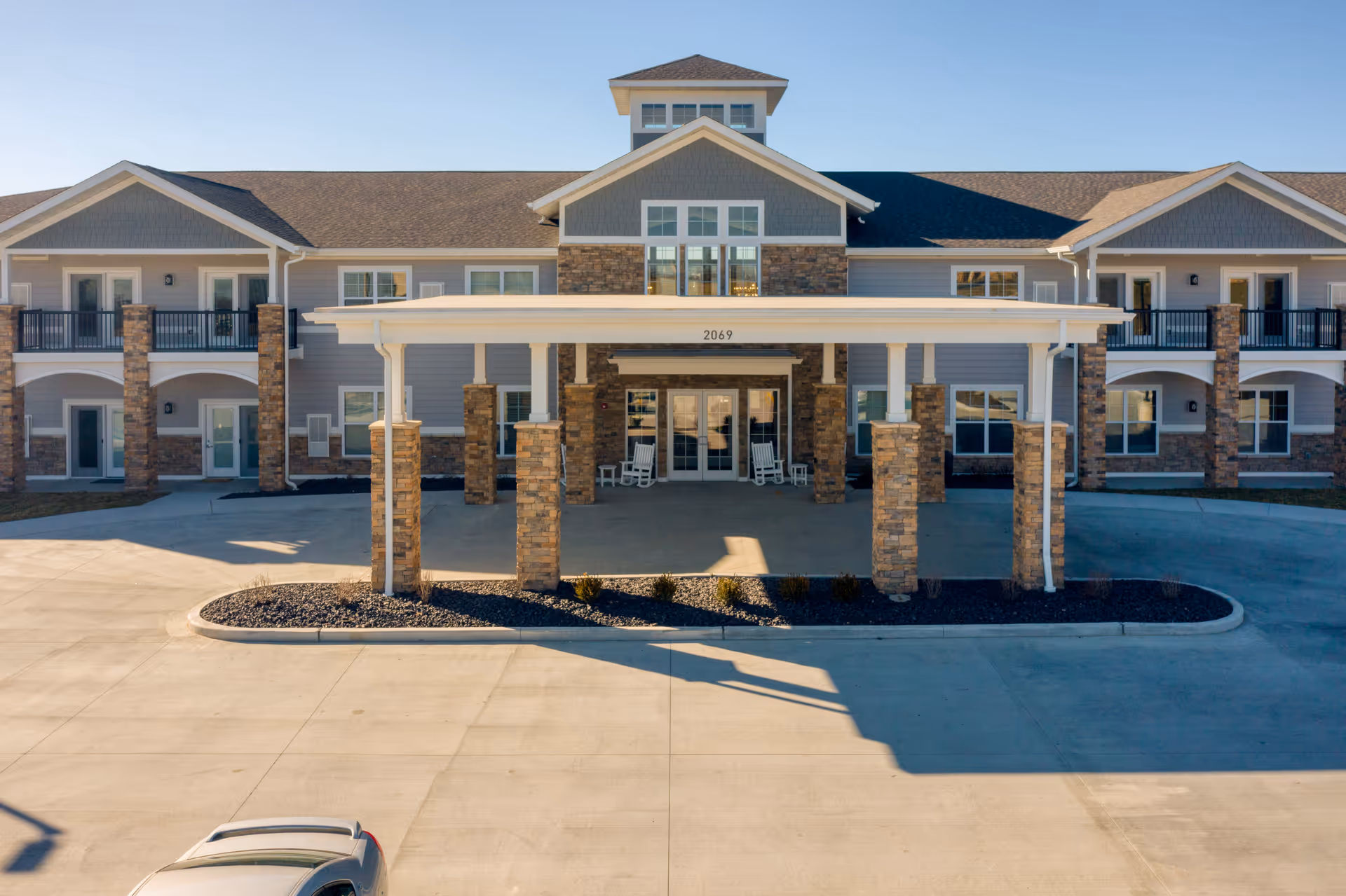 Front exterior view of Cedarhurst Senior Living of Arnold, showing a two-story building with stone pillars, a covered entrance with rocking chairs, and a clear blue sky.
