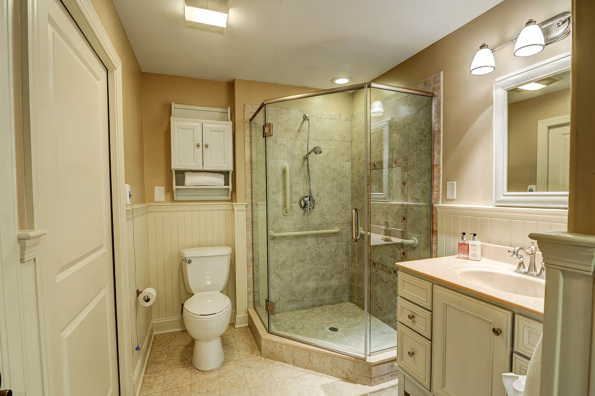 A clean and well-lit bathroom featuring a glass-enclosed corner shower with beige tiled walls and grab bars, a white toilet with a toilet paper holder on the wall, a white cabinet with towels above the toilet, and a white vanity with a sink, faucet, soap dispensers, and a large mirror with two wall-mounted lights above it.