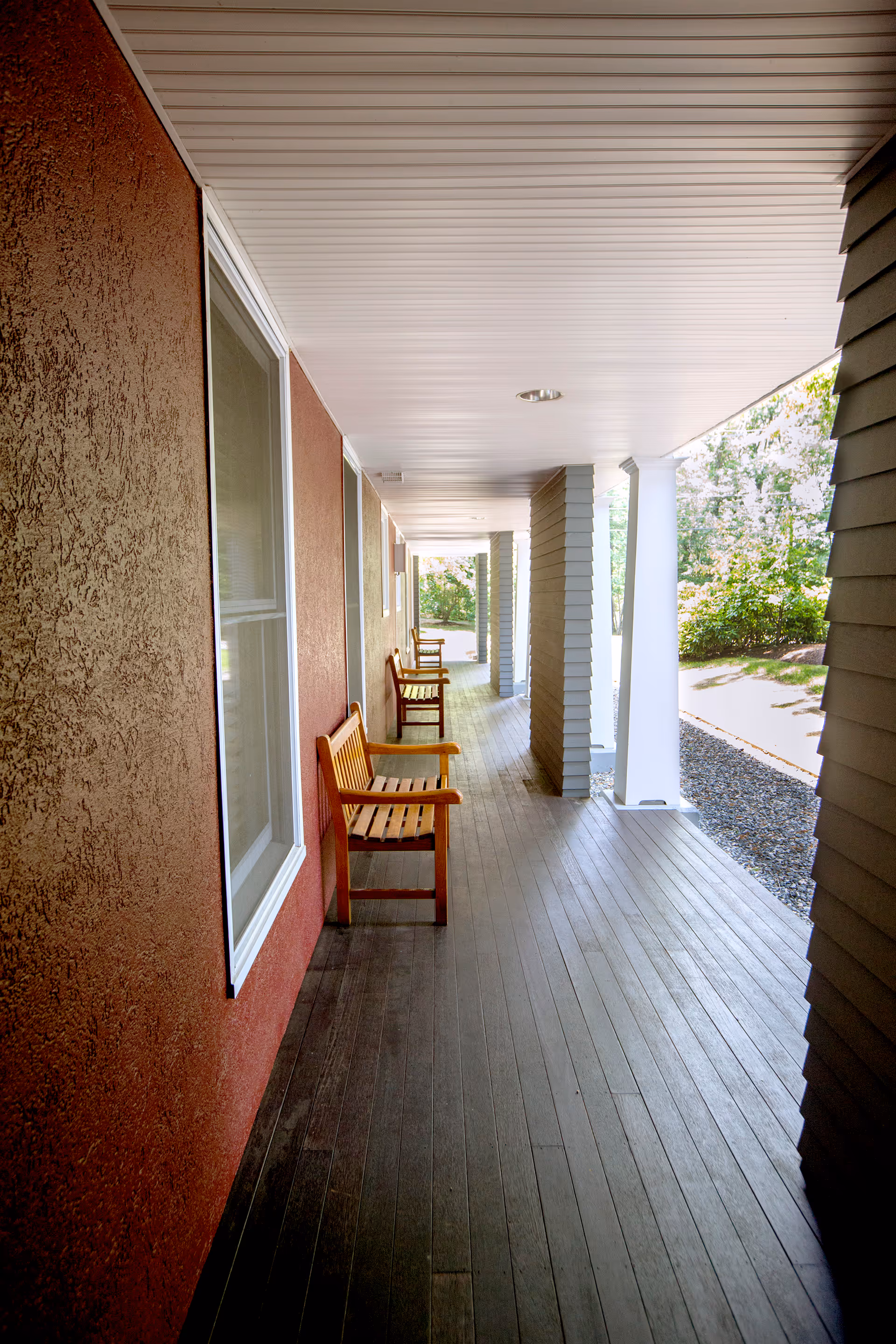 A covered outdoor porch area with wooden benches along a red textured wall, white ceiling, and columns. The porch overlooks a garden area with greenery and trees.