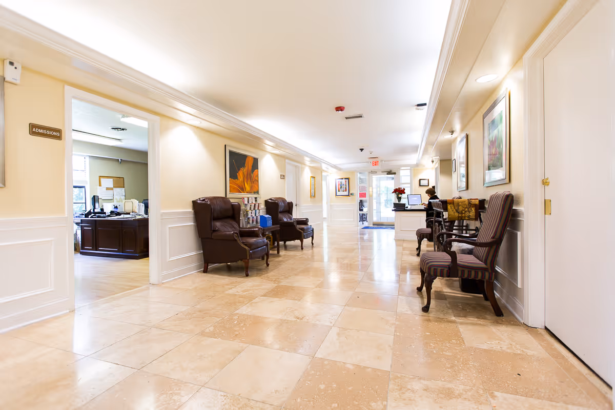 A bright and clean hallway in a senior living facility with beige tiled floors and cream-colored walls. The hallway features several upholstered chairs along the walls, framed artwork, and a reception desk at the far end where a person is seated working on a computer. To the left, there is an open door labeled 'Admissions' leading to an office with desks and computers.