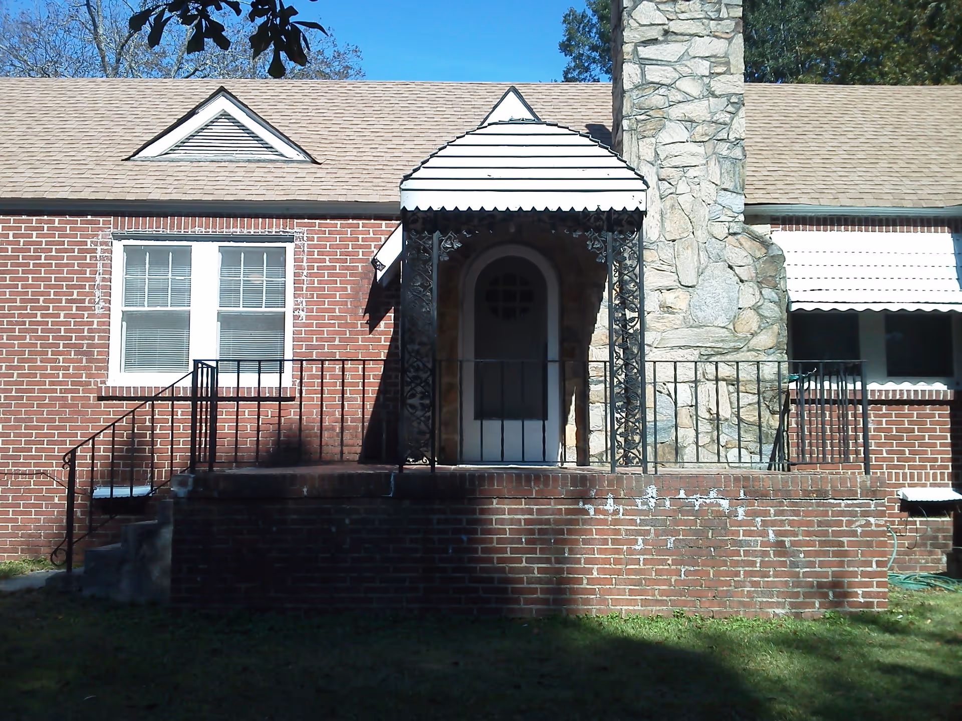 Front exterior view of a single-story brick house with a small covered porch, black wrought iron railings, a stone chimney, and white window frames under a clear blue sky.
