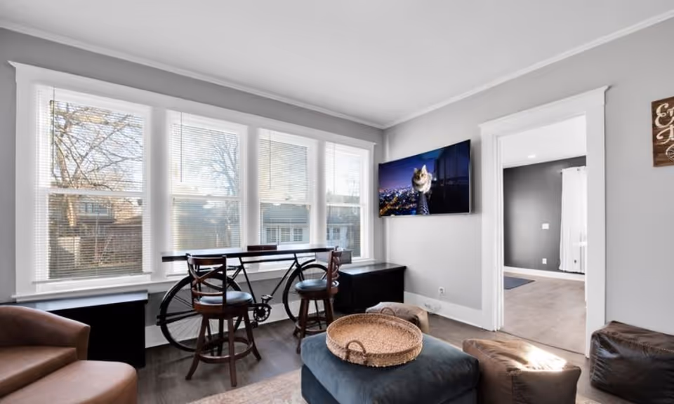Bright living room with large windows, a bar-height table and stools with a bicycle, wall-mounted TV, and seating including an ottoman and chairs.