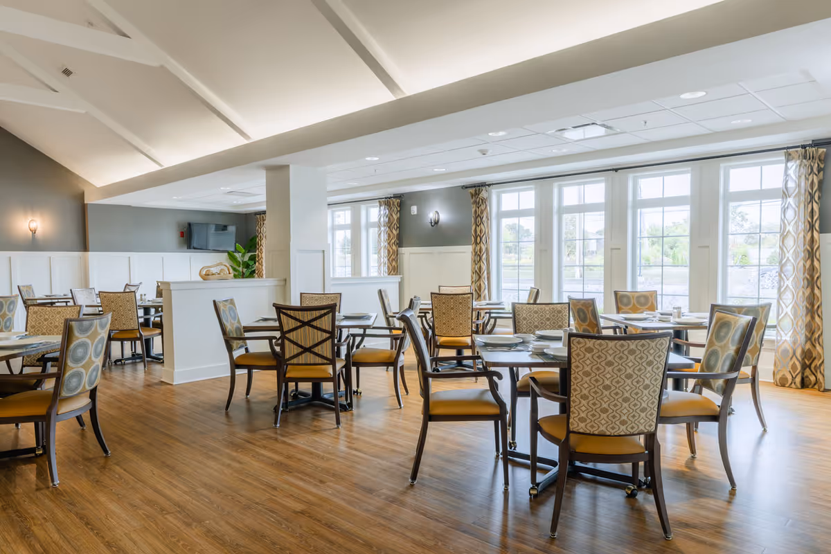 A bright and spacious dining room with multiple tables and chairs arranged neatly. The room features large windows with patterned curtains allowing natural light to fill the space. The floor is wooden, and the walls are painted in neutral tones with white paneling. There is a TV mounted on one wall and a decorative plant near the corner.