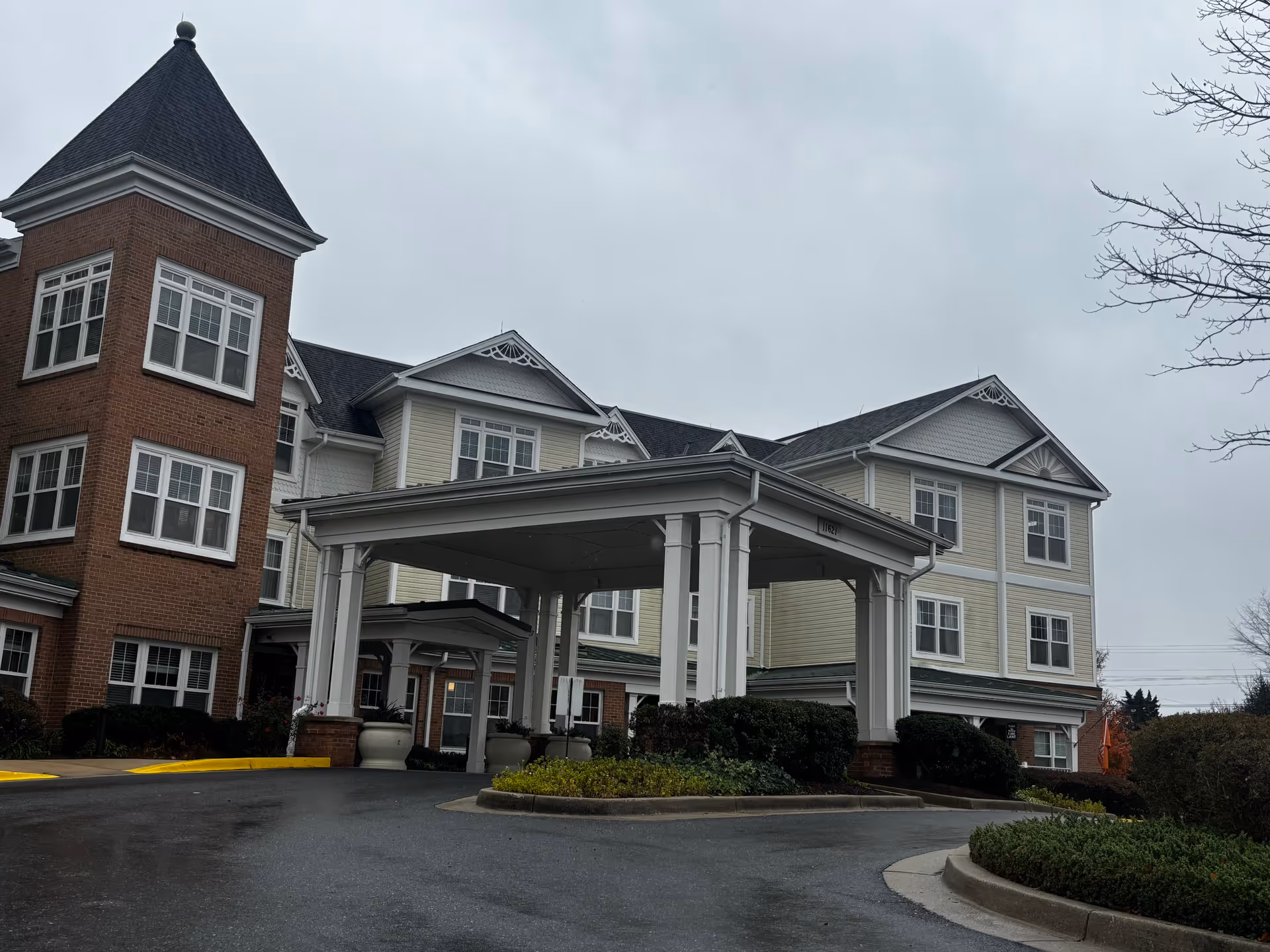 Exterior view of a senior living facility building with a covered entrance driveway, brick and light-colored siding, multiple windows, and a cloudy sky.