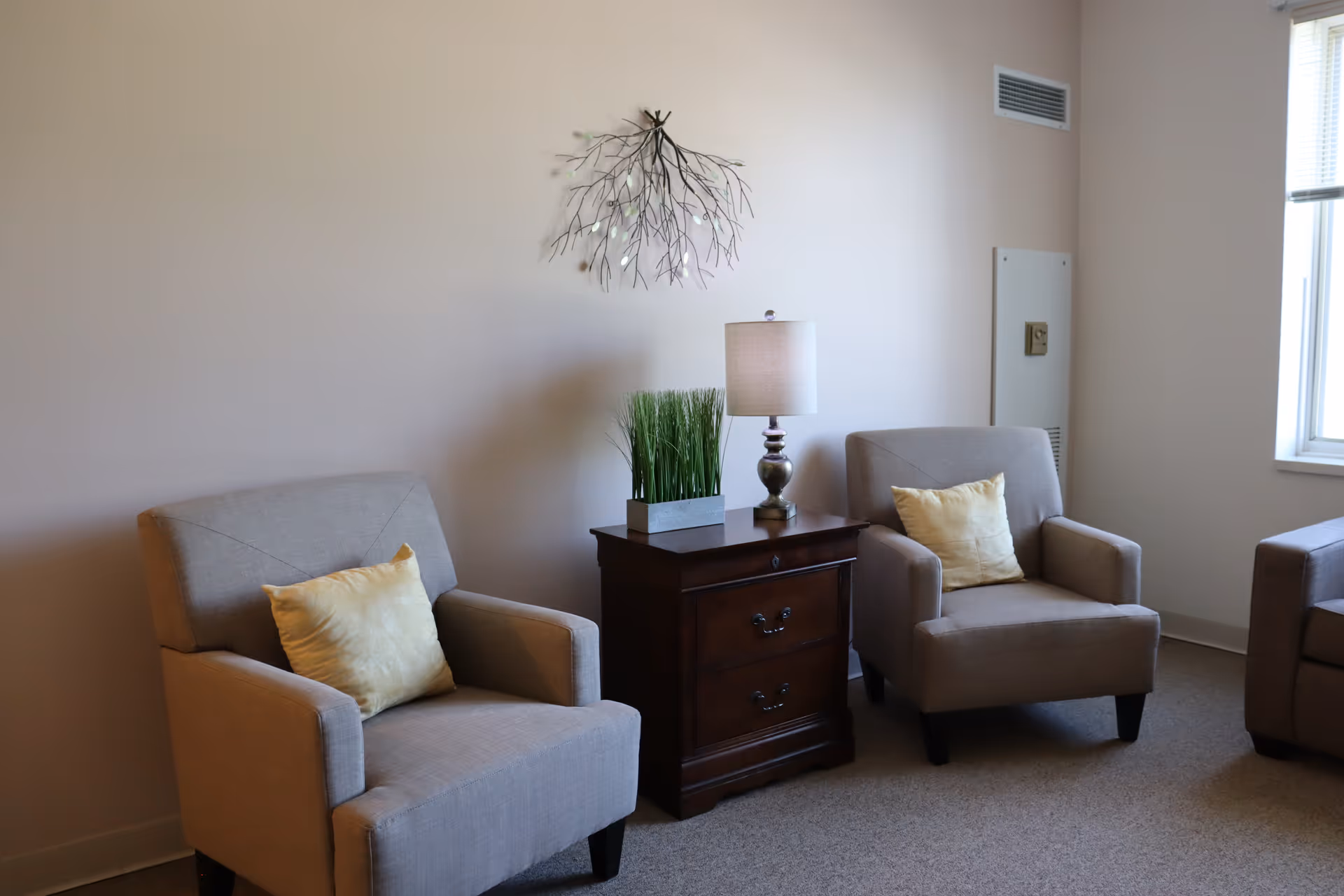 A cozy sitting area with two beige armchairs each with a yellow cushion, separated by a dark wooden side table holding a lamp and a rectangular planter with green grass. A decorative metal wall art piece resembling branches is mounted on the light-colored wall behind the chairs. A window with blinds is partially visible on the right side.