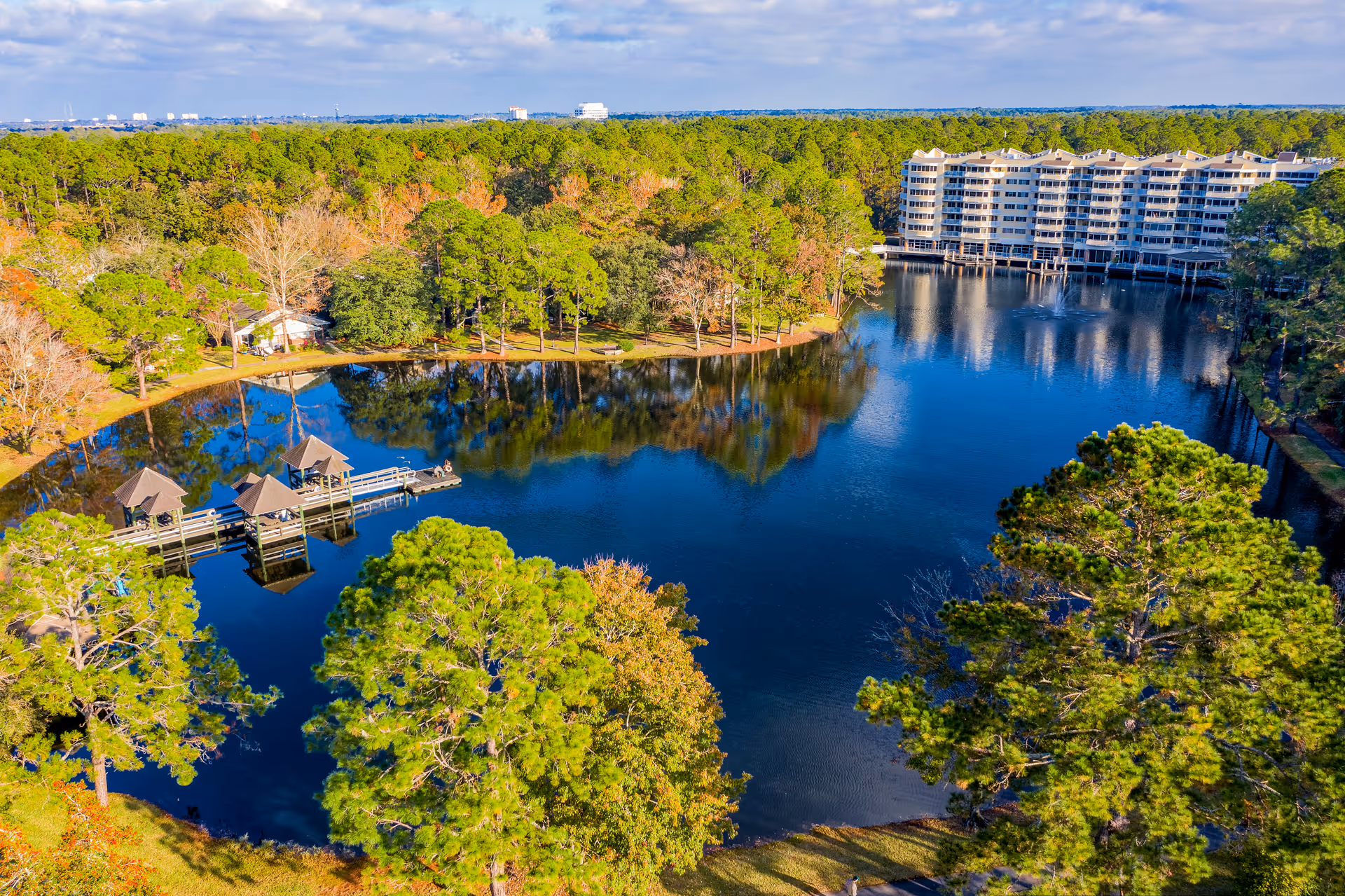 Aerial view of a large pond surrounded by trees with a multi-story residential building on the far side. The pond has a dock with small covered seating areas extending over the water. The surrounding area is lush with green and autumn-colored trees under a clear blue sky.