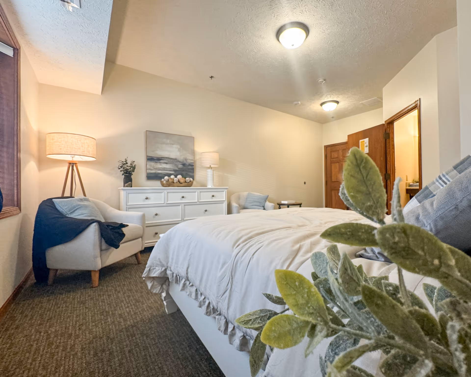 A cozy bedroom with a large bed covered in white bedding, a white dresser with decorative items and a painting above it, two armchairs with a blanket draped over one, a floor lamp, and a plant in the foreground. The room has beige walls, carpeted floor, and wooden doors.