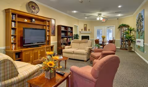 A cozy senior living facility common area with comfortable upholstered chairs and sofas arranged around wooden coffee tables. There is a large wooden entertainment center with a flat-screen TV, bookshelves, and decorative items. The room is warmly lit with ceiling lights and a ceiling fan, and there are plants and artwork decorating the space. In the background, there is a dining area with a table and chairs near glass doors.