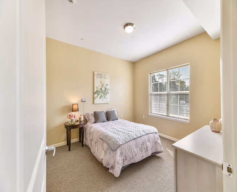 Small furnished bedroom with a bed in floral bedding, a nightstand with lamp, a dresser, and a window letting in daylight.