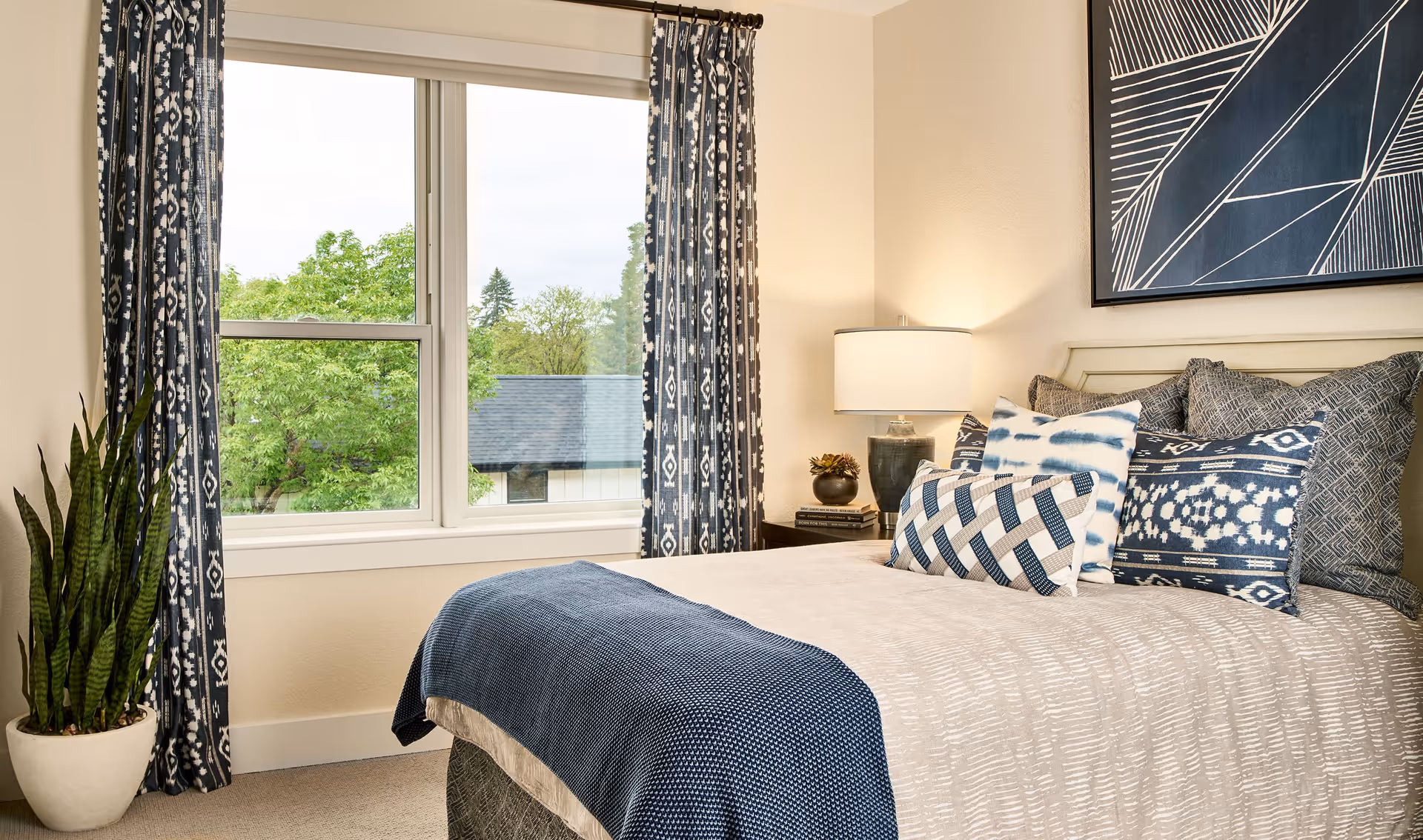 A neatly made bedroom with blue-and-white bedding, patterned pillows, a bedside lamp, potted plant, and a window with patterned curtains overlooking trees.