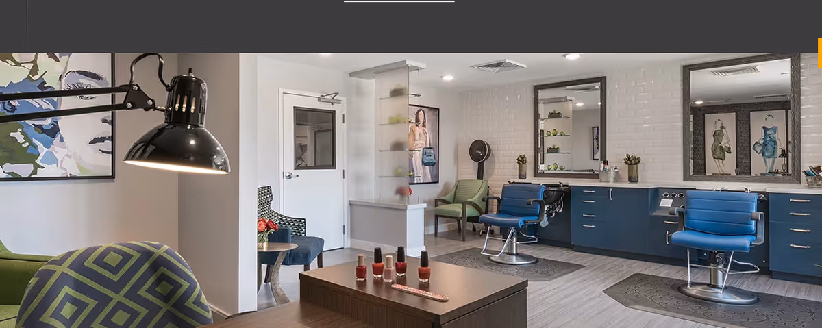Interior view of a salon area in a senior living facility with two blue salon chairs in front of large mirrors, a hair dryer, green and blue patterned chairs, a table with nail polish bottles, and modern decor including artwork on the walls.