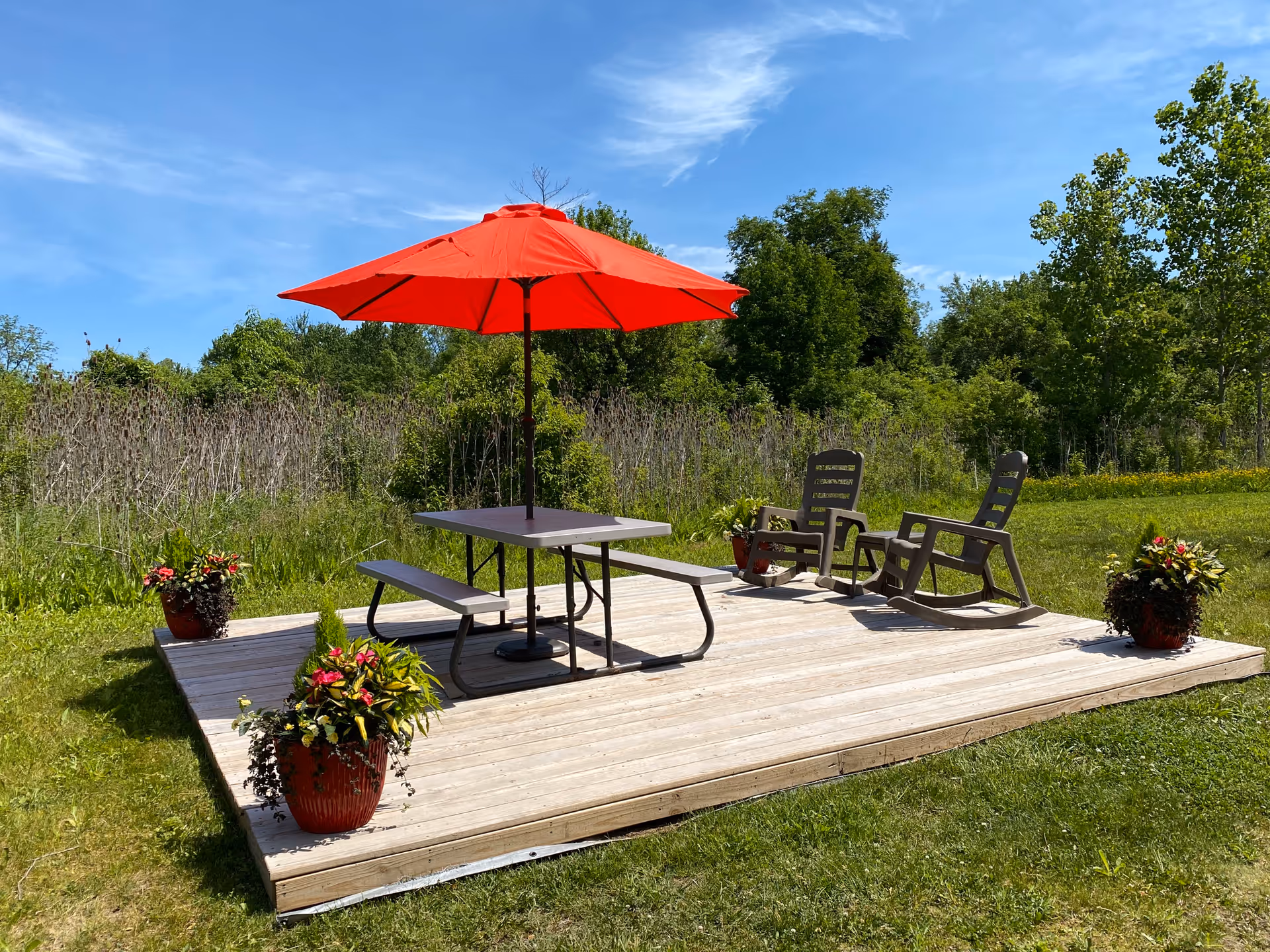 Outdoor wooden deck with a picnic table and benches under a large red umbrella, two rocking chairs, and four potted plants with flowers, surrounded by green grass and trees under a blue sky.