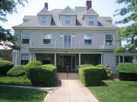 Two-story gray senior living building with a covered front porch and manicured shrubs.