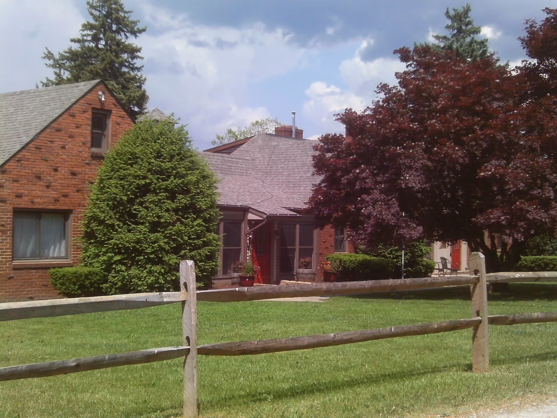 Exterior view of a brick building with a shingled roof, surrounded by green grass, a wooden fence, and various trees including a large green bush and a red-leafed tree under a partly cloudy sky.