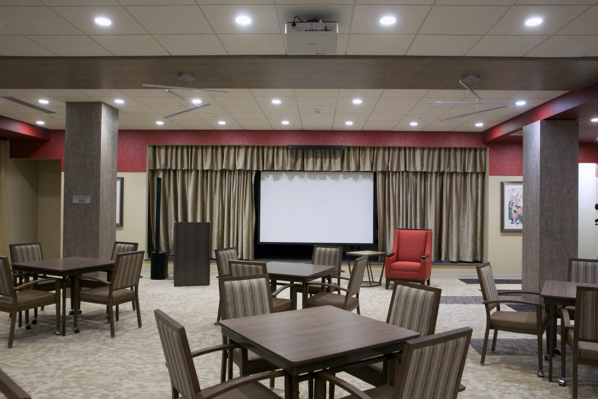 A spacious room with multiple square tables and chairs arranged for group seating. At the far end, there is a large white projection screen with beige curtains on either side. A red armchair and a small table are positioned near the screen. The room has a beige carpet with a subtle pattern, and the ceiling is fitted with recessed lighting and a projector.