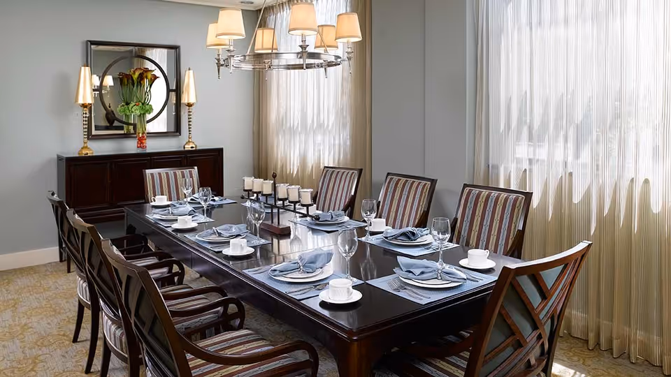 A formal dining room with a long dark wooden table set for eight people. Each place setting includes a plate, napkin, glass, and cup. The chairs have striped upholstery. A sideboard with two lamps and a vase of flowers is against the wall, and a round mirror hangs above it. Sheer curtains cover the windows, and a chandelier with multiple lampshades hangs above the table.