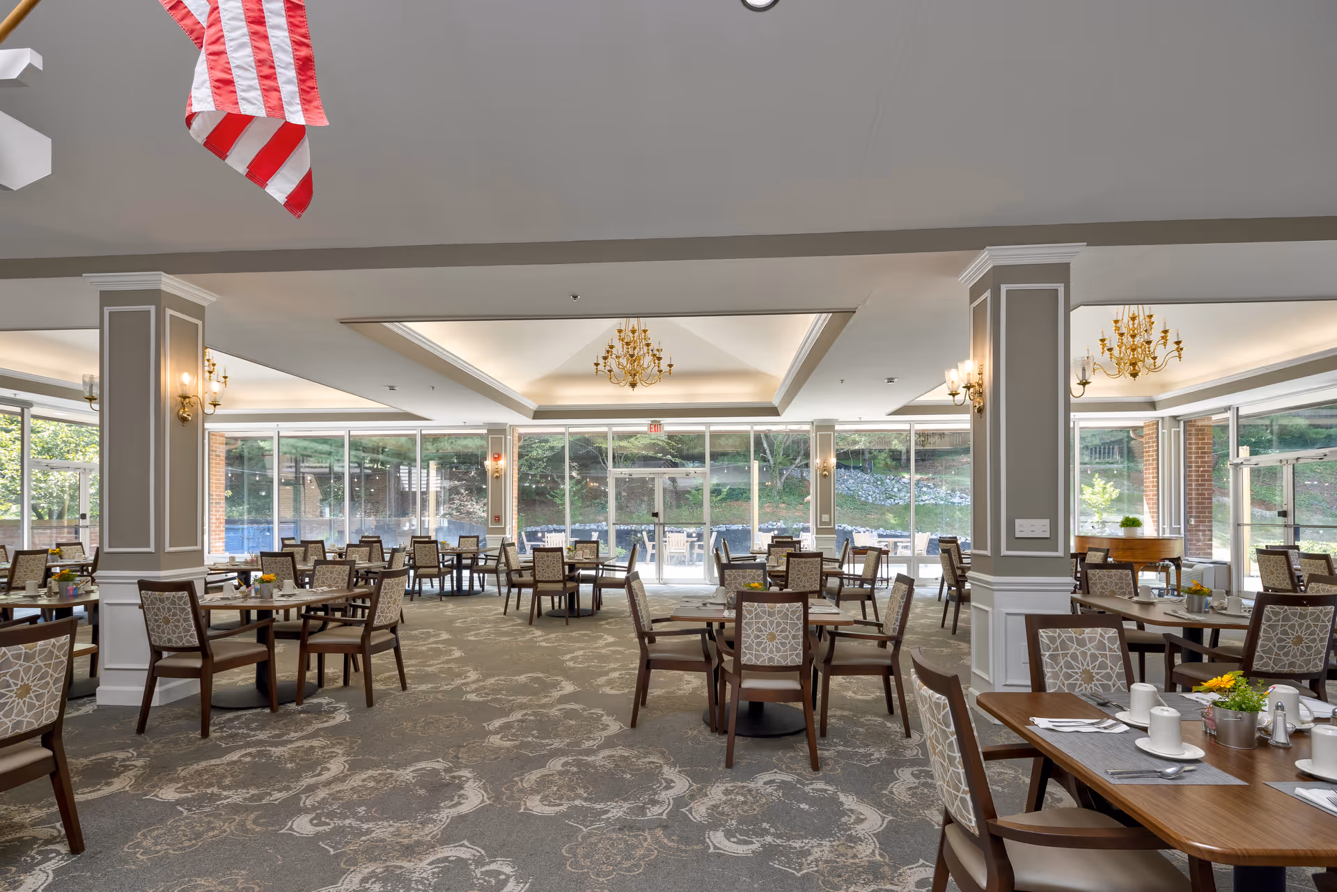 Bright dining room with multiple tables and upholstered chairs, chandeliers, and large floor-to-ceiling windows overlooking greenery.