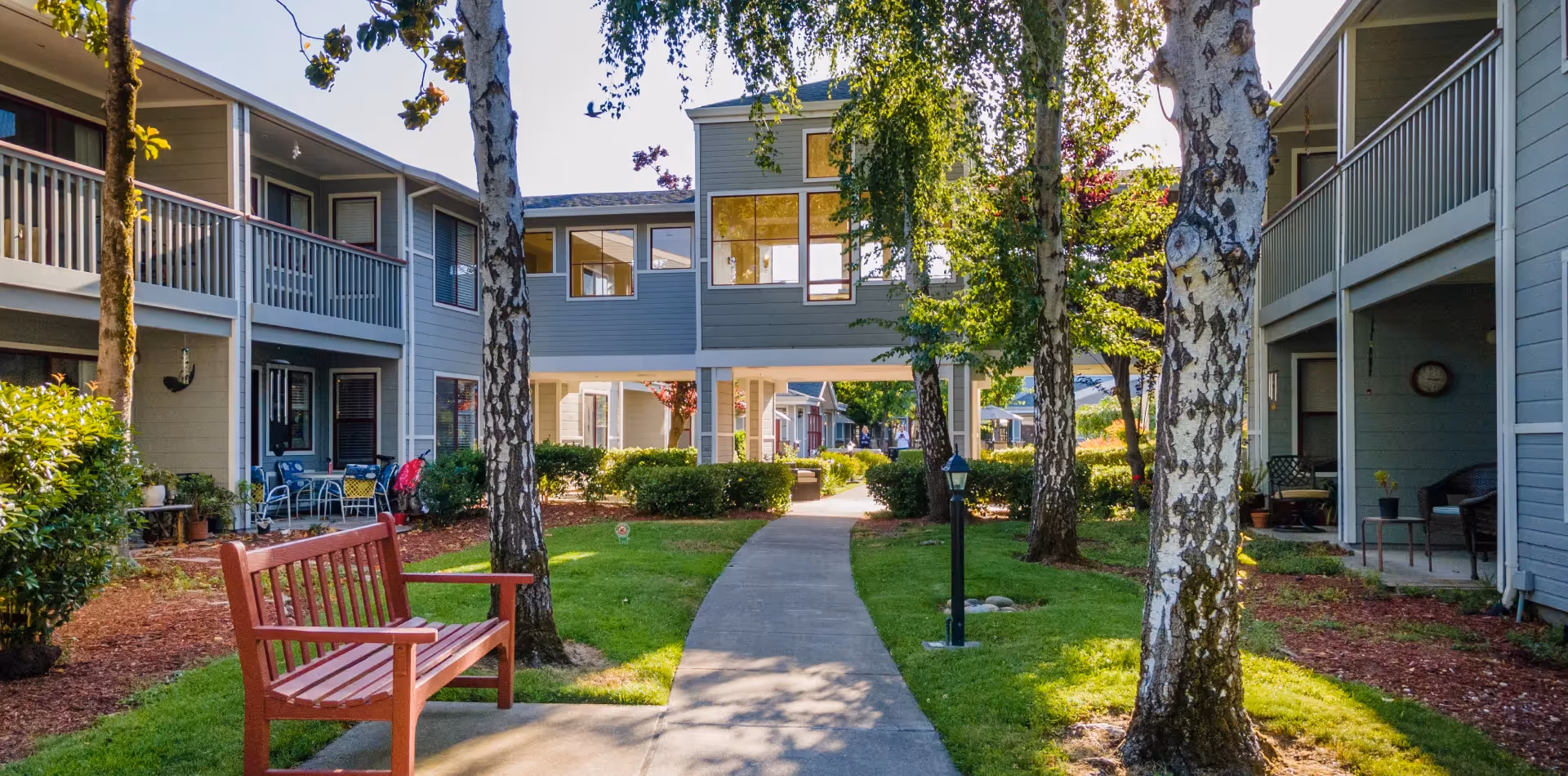 Outdoor courtyard area of a senior living facility with a paved walkway, green grass, trees, and a wooden bench. The buildings surrounding the courtyard have balconies and patios with outdoor furniture.