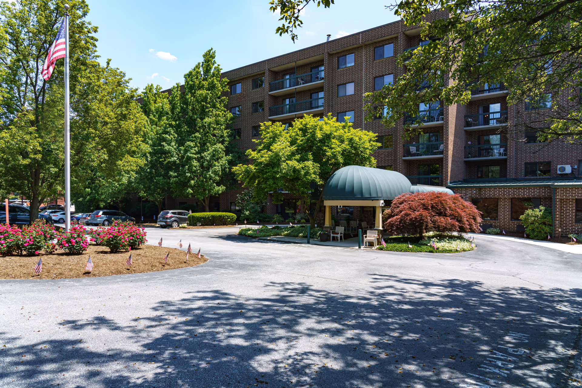Front entrance of a multi-story brick senior living building with a covered drive-through canopy, balconies, landscaped roundabout and an American flag.