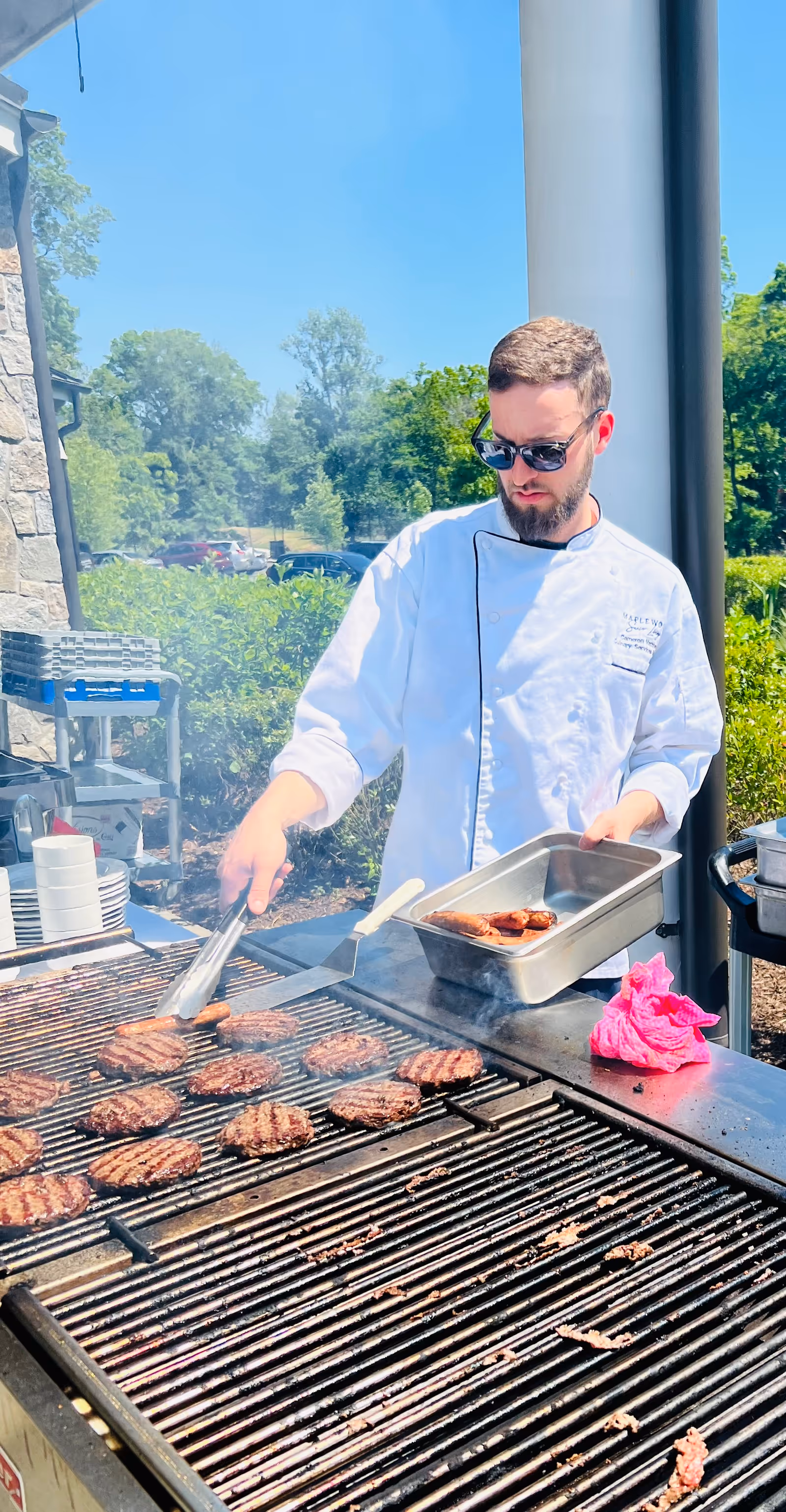 A man wearing sunglasses and a white chef's coat is grilling multiple hamburger patties on a large outdoor grill. He is holding a metal tray with cooked patties in one hand and using tongs to handle the patties on the grill. The background shows greenery, trees, and a clear blue sky.