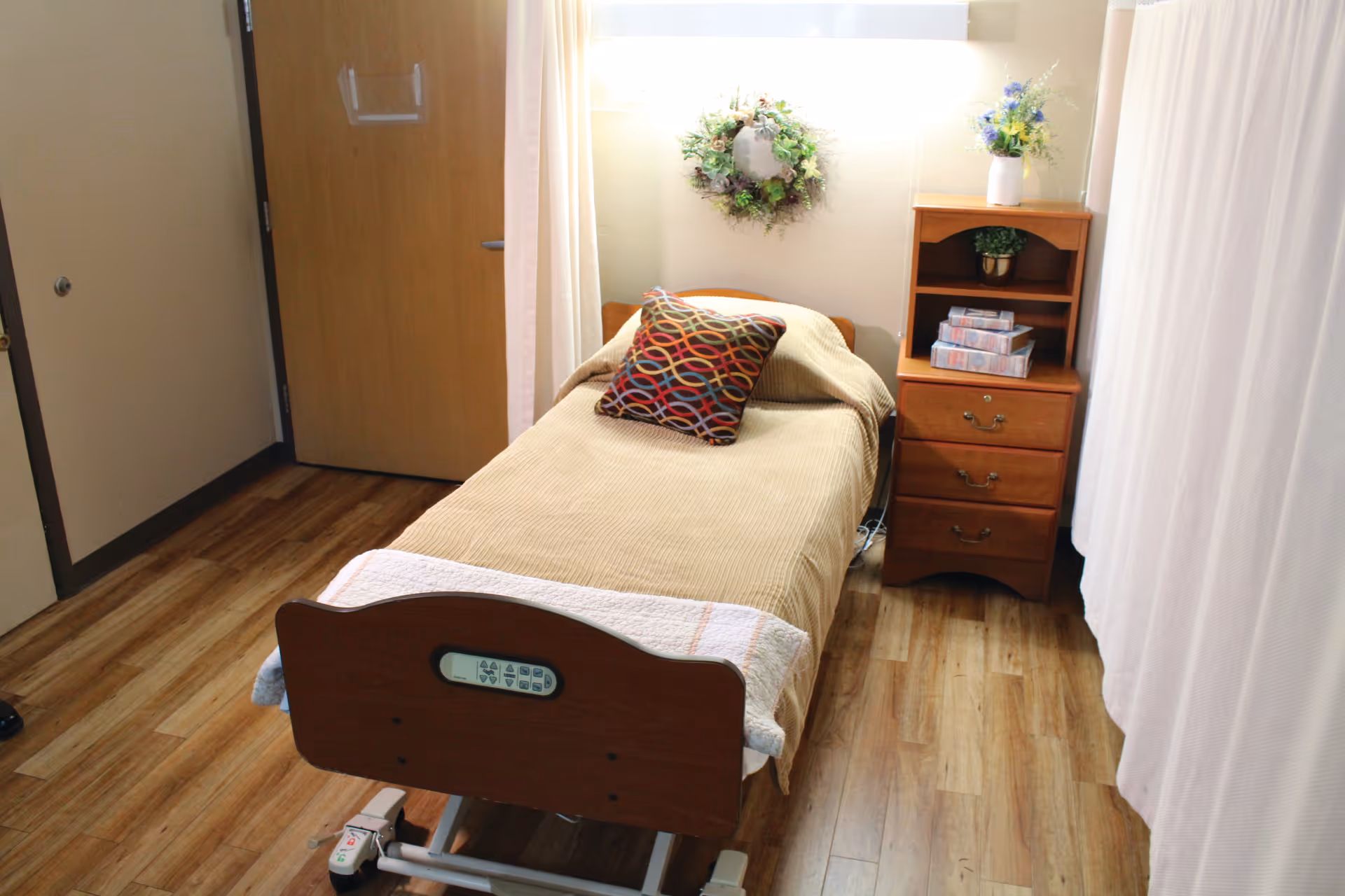 Single hospital-style bed in a small furnished patient room with a wooden nightstand, decorative wreath, and hardwood floor.