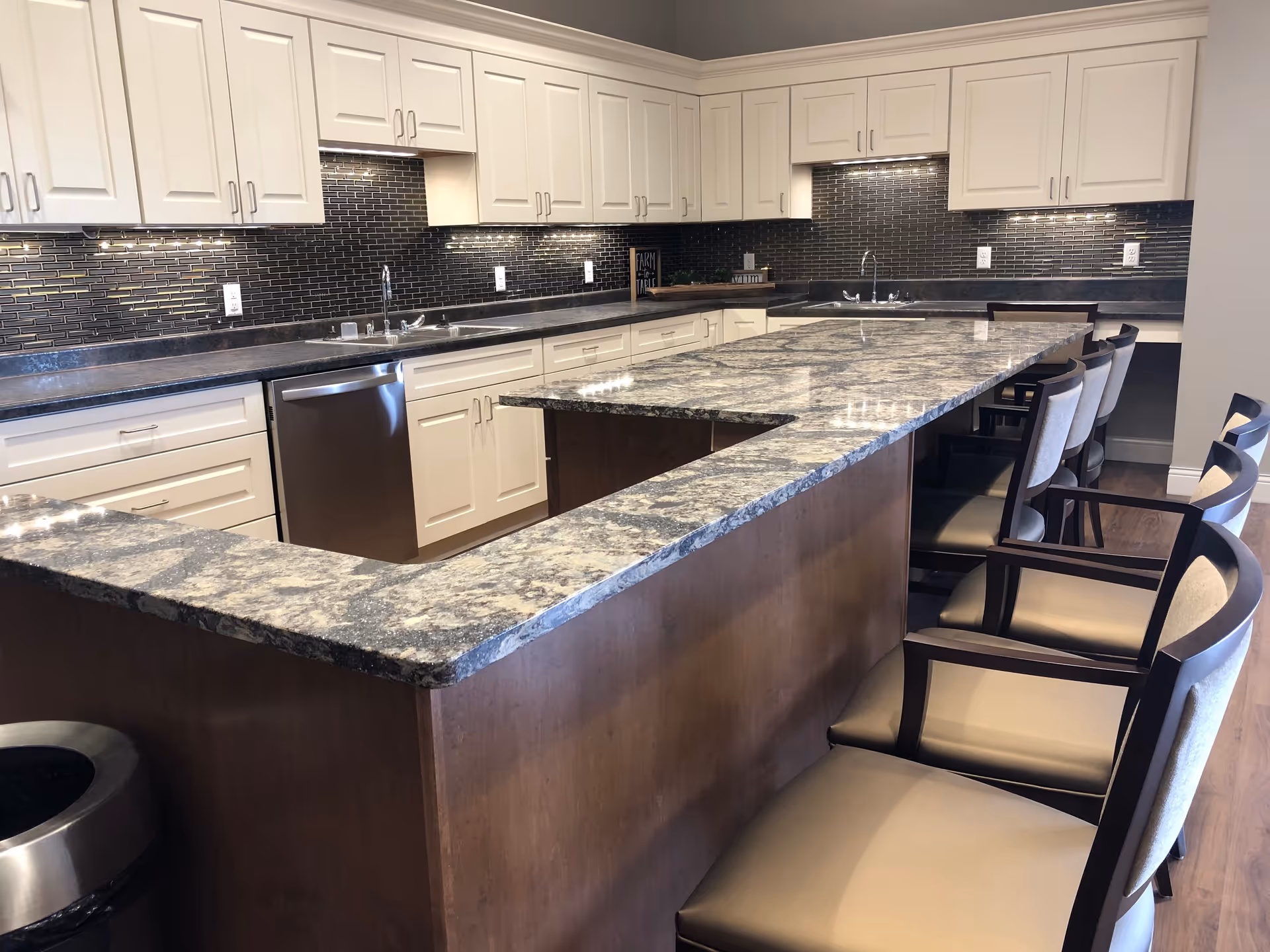 Communal kitchen featuring a long granite island with bar stools, white cabinetry, a stainless dishwasher, and a dark tiled backsplash.