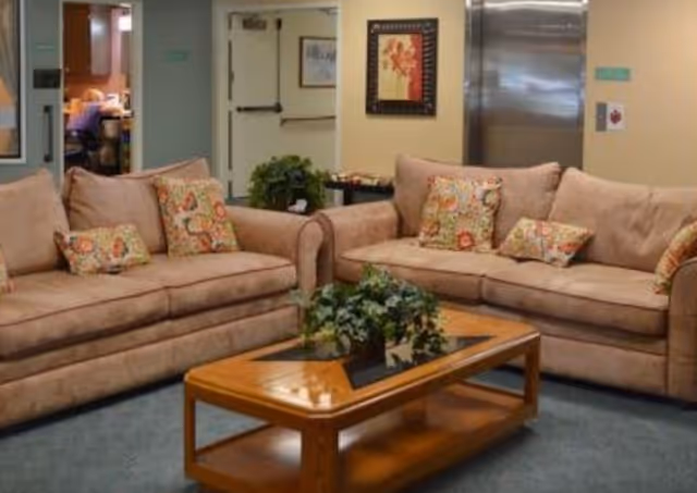 A cozy living room area with two beige sofas adorned with colorful floral patterned pillows. In front of the sofas is a wooden coffee table with a glass inset and a green potted plant on top. The background shows a hallway with a door and framed artwork on the wall.