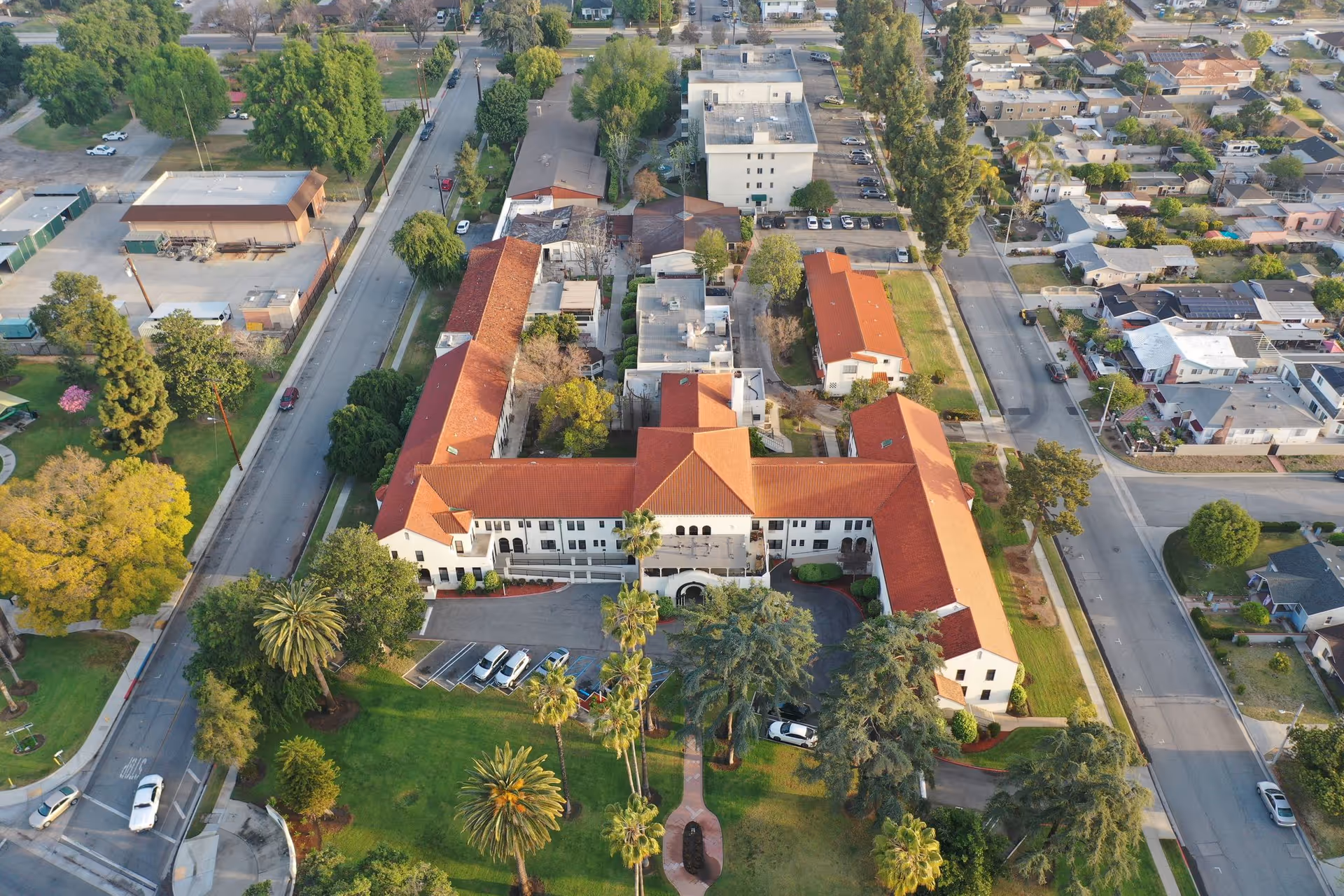 Aerial view of the California Mission Inn facility showing a large building with red tile roofing surrounded by green lawns, palm trees, and other trees. The building is situated in a residential neighborhood with streets and houses visible around it.