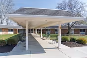 Covered walkway entrance to a single-story brick building with shrubs and trees in the background under a clear sky.