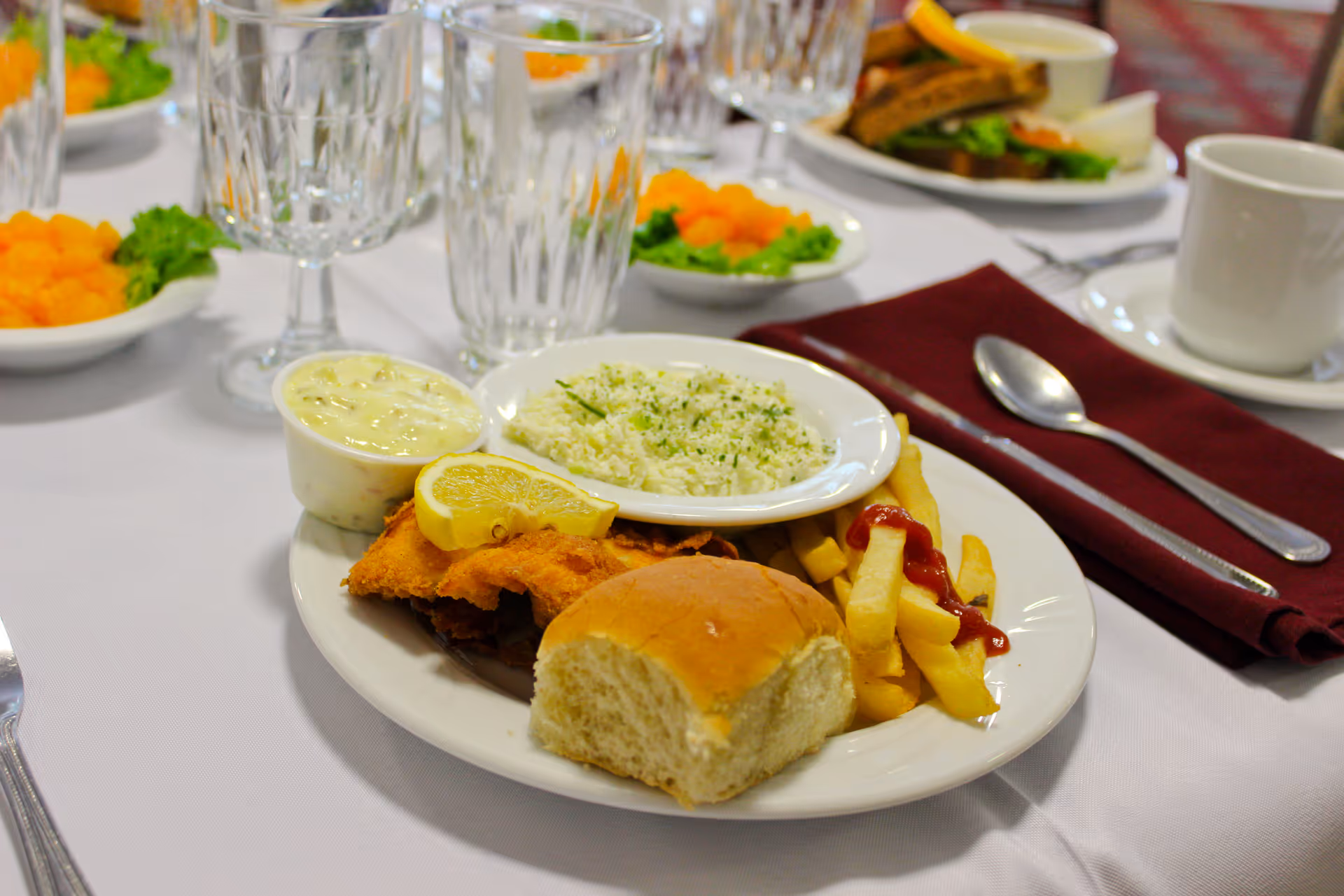 A plated meal on a white tablecloth featuring fried fish with a lemon wedge, a dinner roll, French fries with ketchup, a small bowl of tartar sauce, and a side of rice garnished with herbs. In the background, there are glasses, a cup, a maroon napkin with a spoon and knife, and other plates with food including salad and sandwiches.