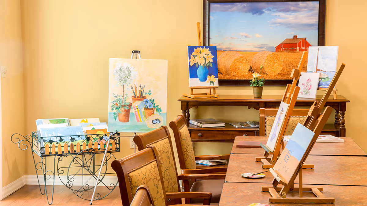 A well-lit art room with several wooden easels holding paintings in progress, a table with chairs, a metal rack holding additional artwork, and a wooden side table with a potted plant and a large framed picture of hay bales and a red barn on the wall.